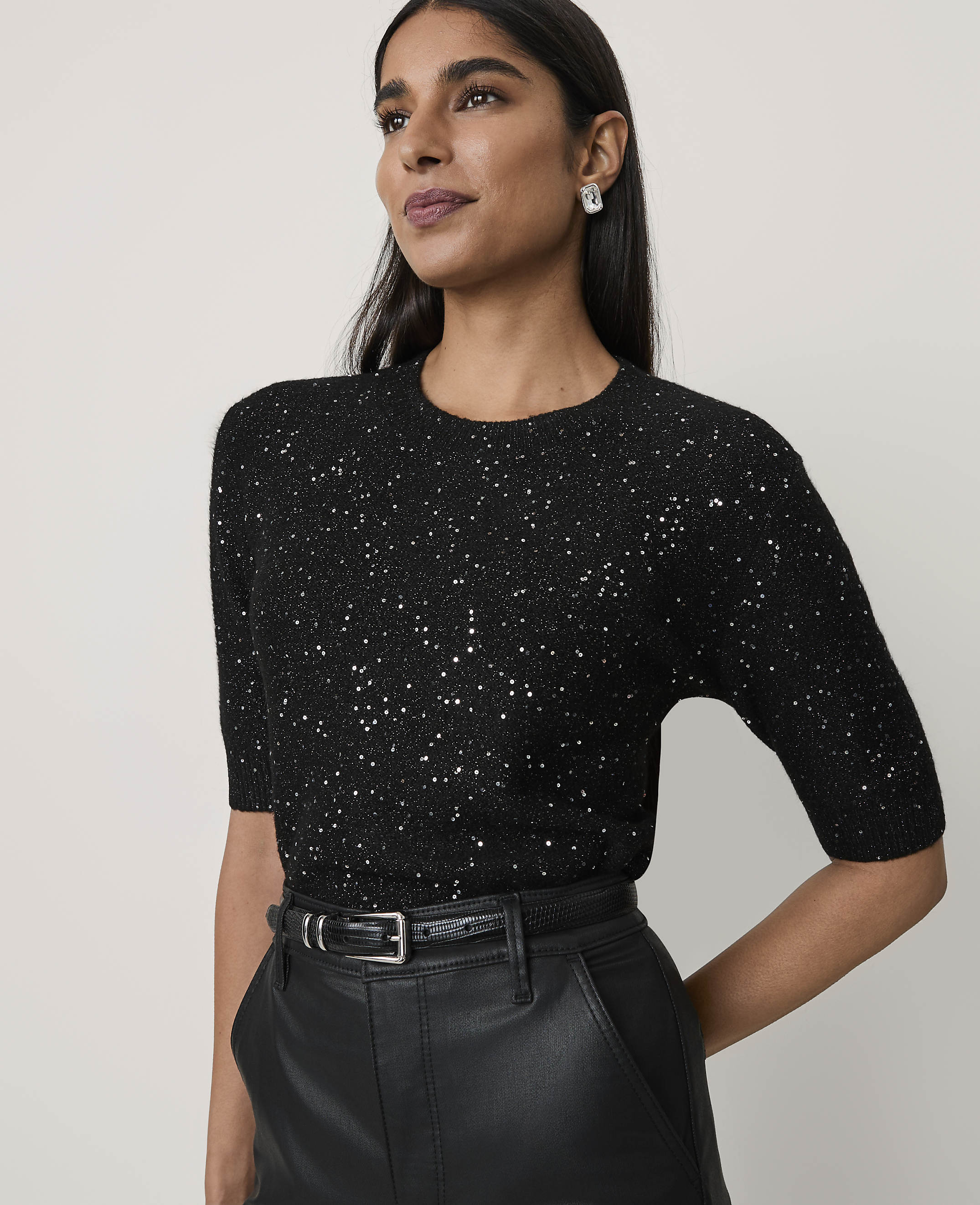 A woman wearing a sparkling black top and leather trousers stands against a plain, light-colored background.