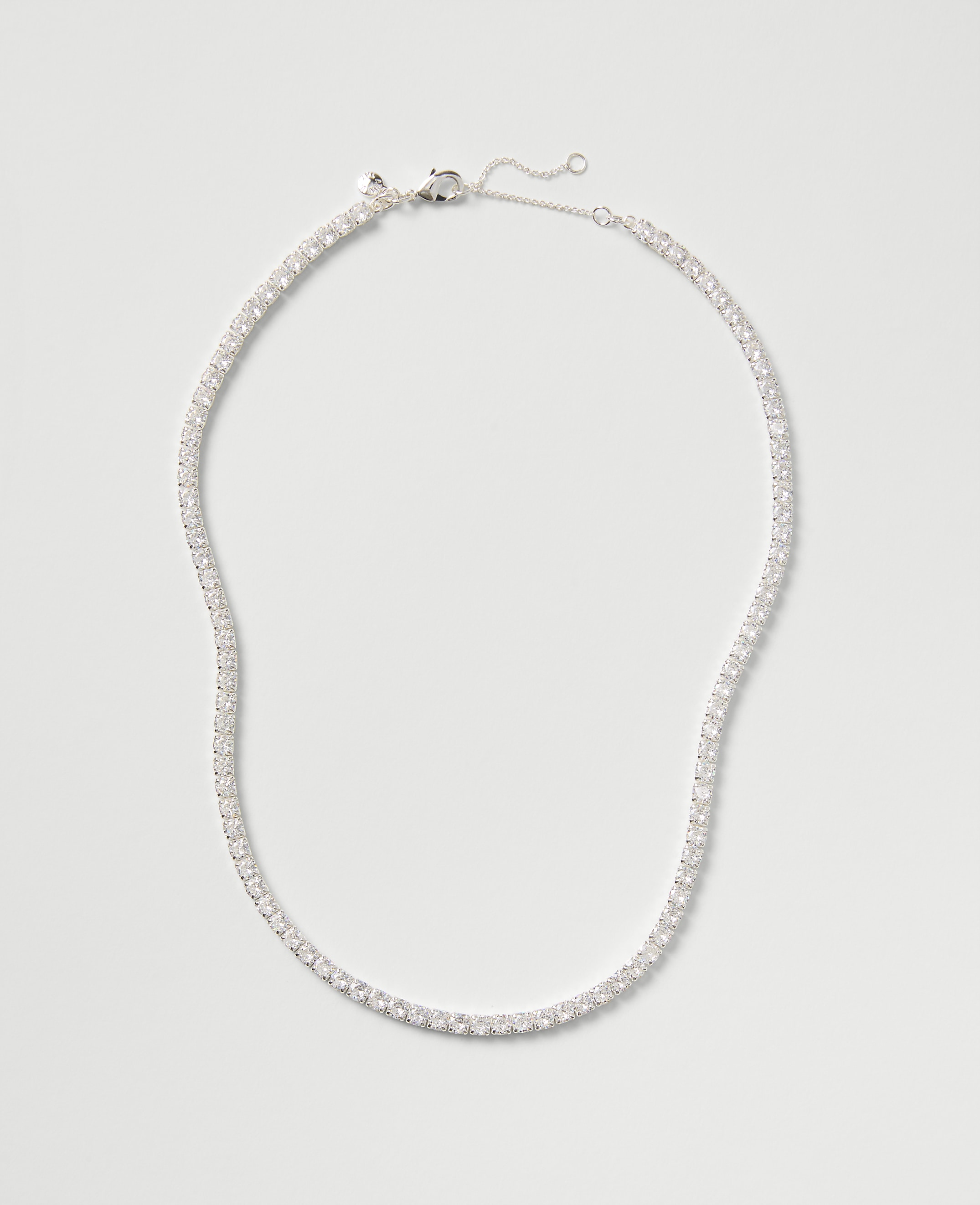 A sparkling tennis necklace with clear stones rests against a plain, light gray background.