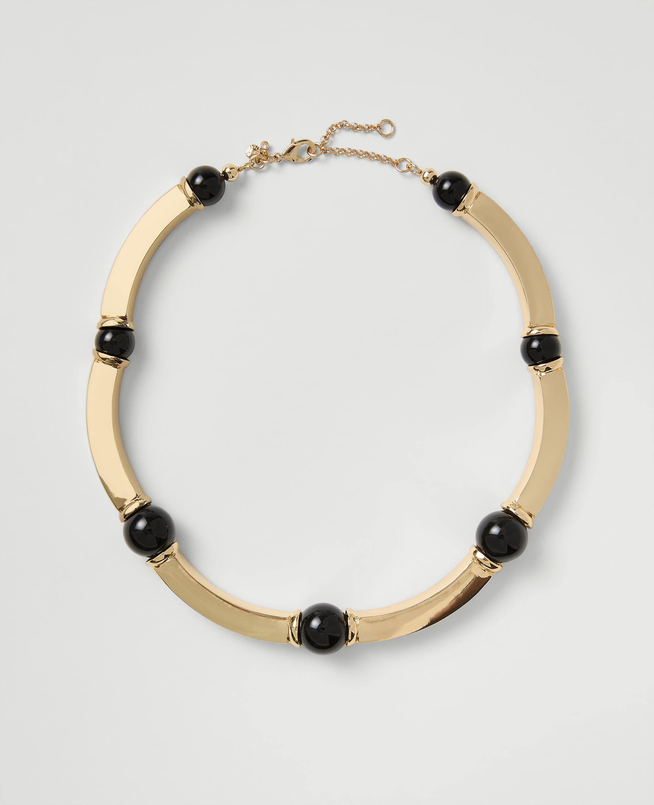 A gold-toned necklace with black spherical beads rests on a plain light gray background.