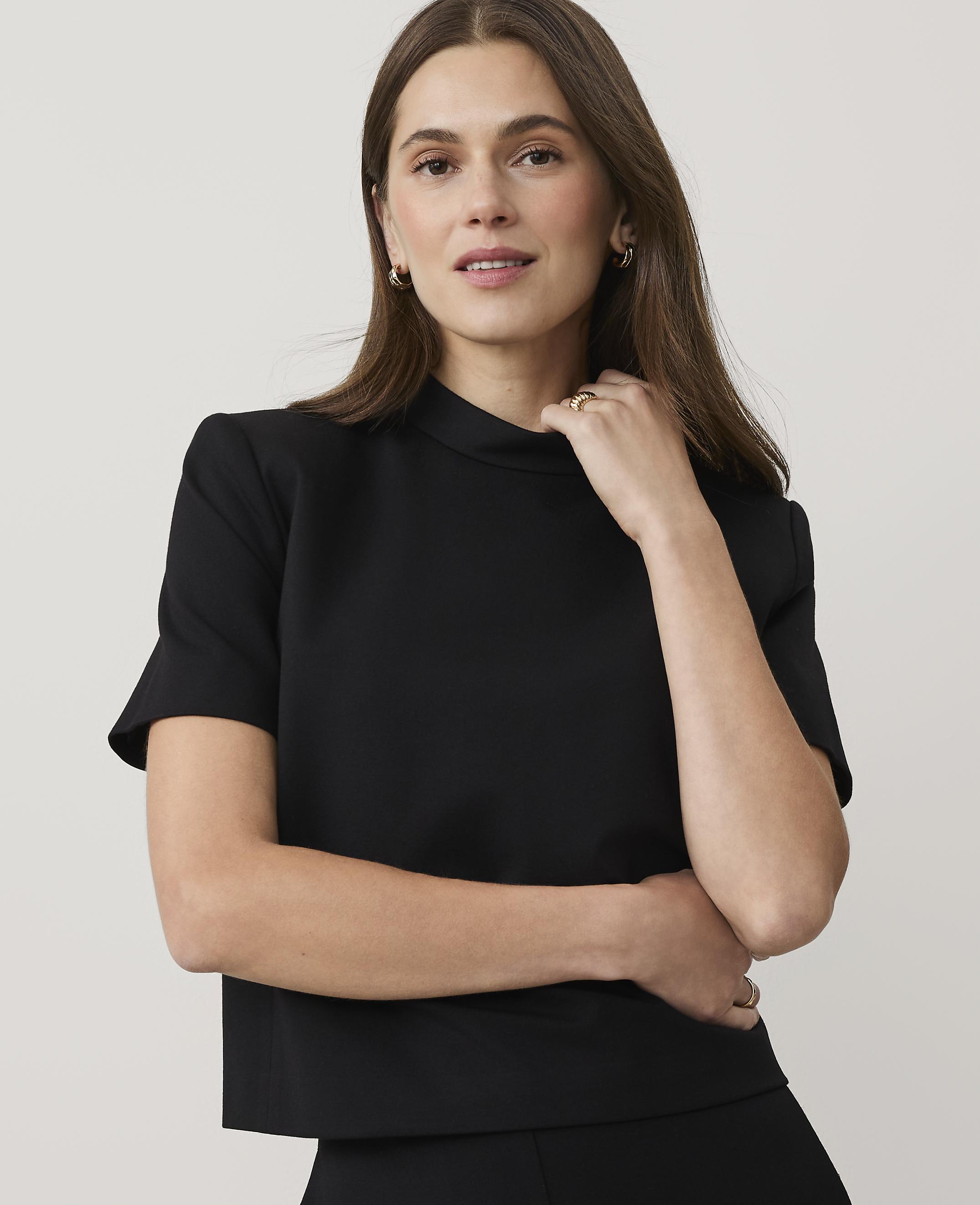 A woman wearing a black short-sleeved top and gold jewelry poses against a plain, light-colored background.