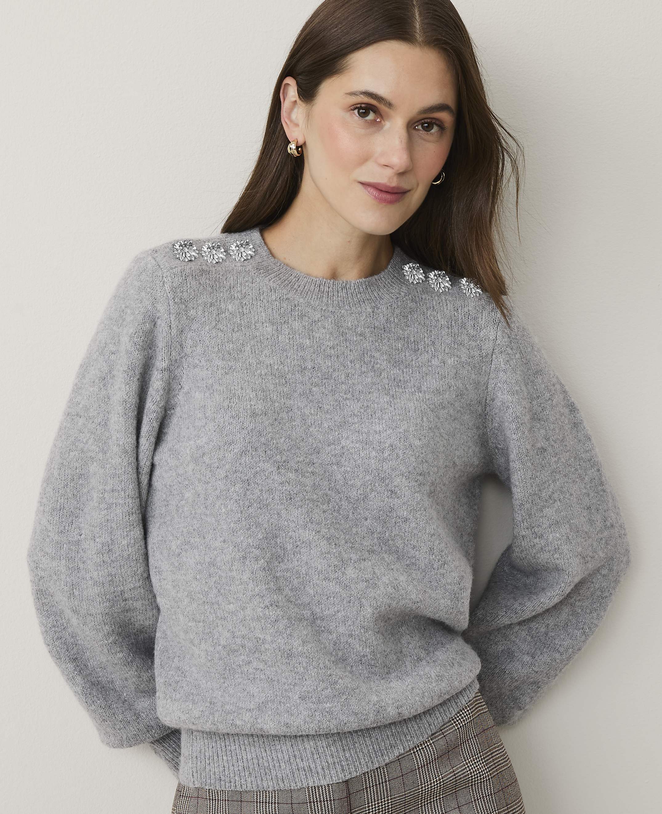 A woman wearing a grey sweater with crystal embellishments on the shoulders stands against a plain, light-colored wall.
