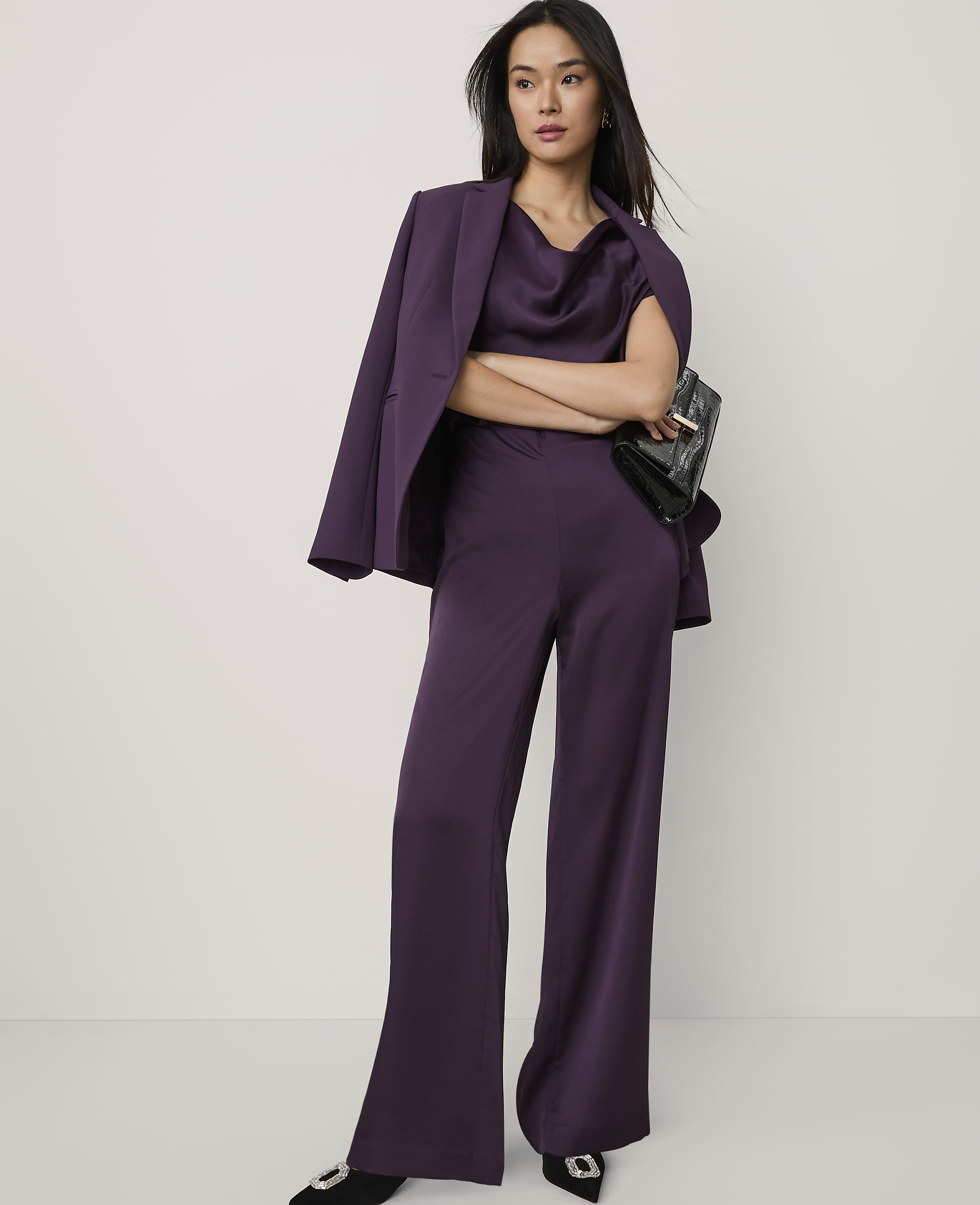 A woman wearing a purple satin suit and holding a black clutch stands against a plain white background.