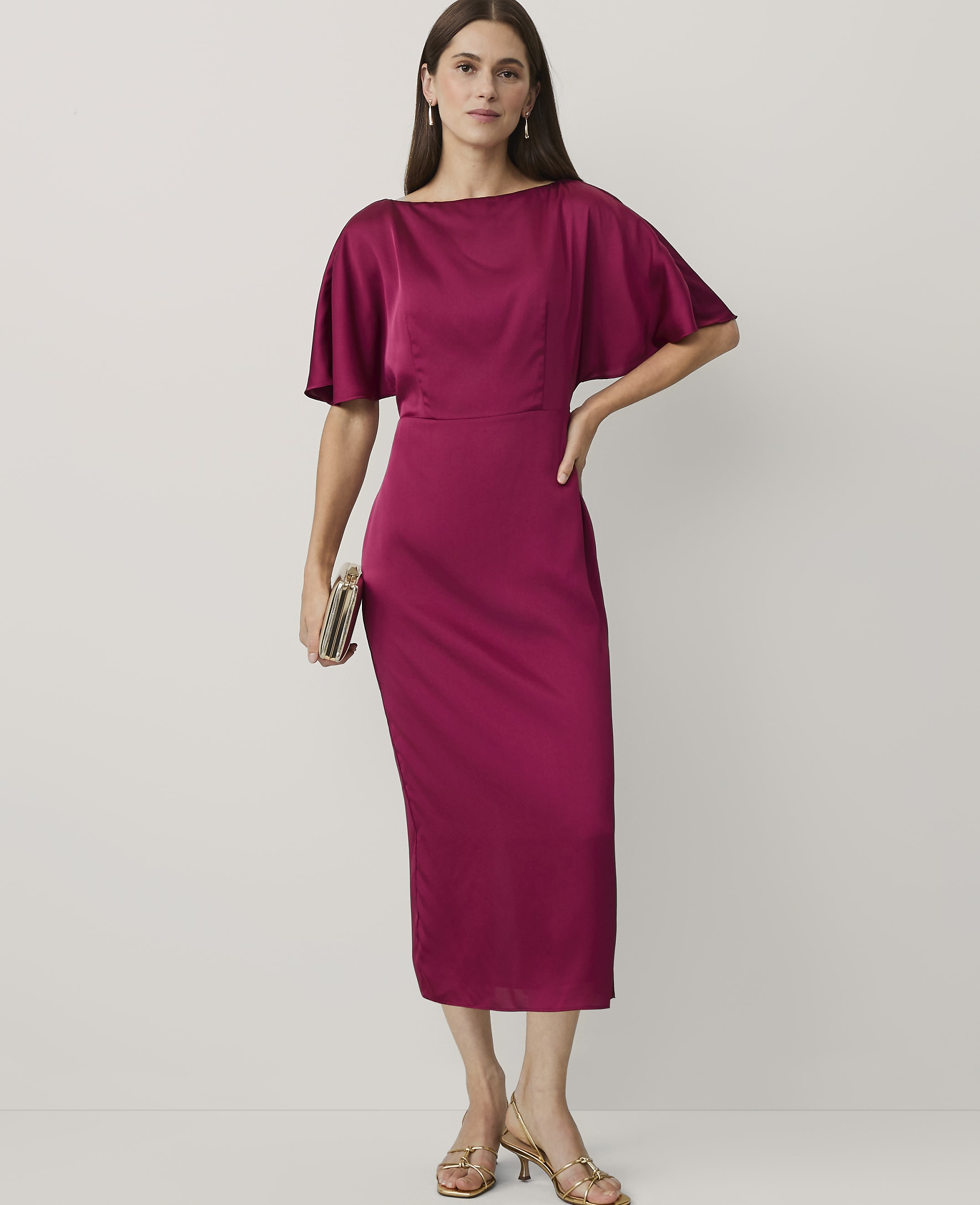 A woman wearing a magenta satin midi dress and gold heels stands against a plain white background.