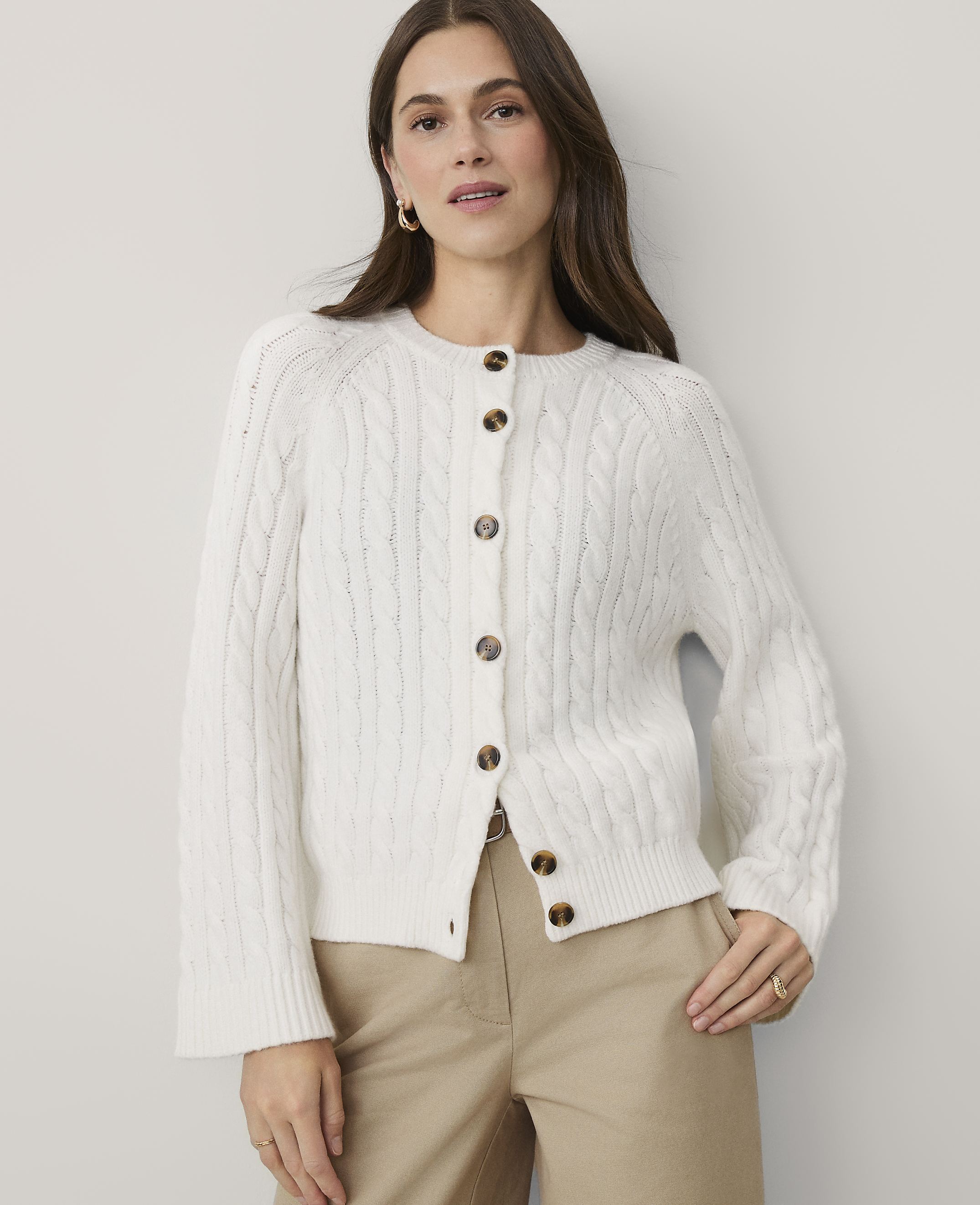 A woman wearing a white cable-knit cardigan and khaki pants stands against a plain, light-colored wall.