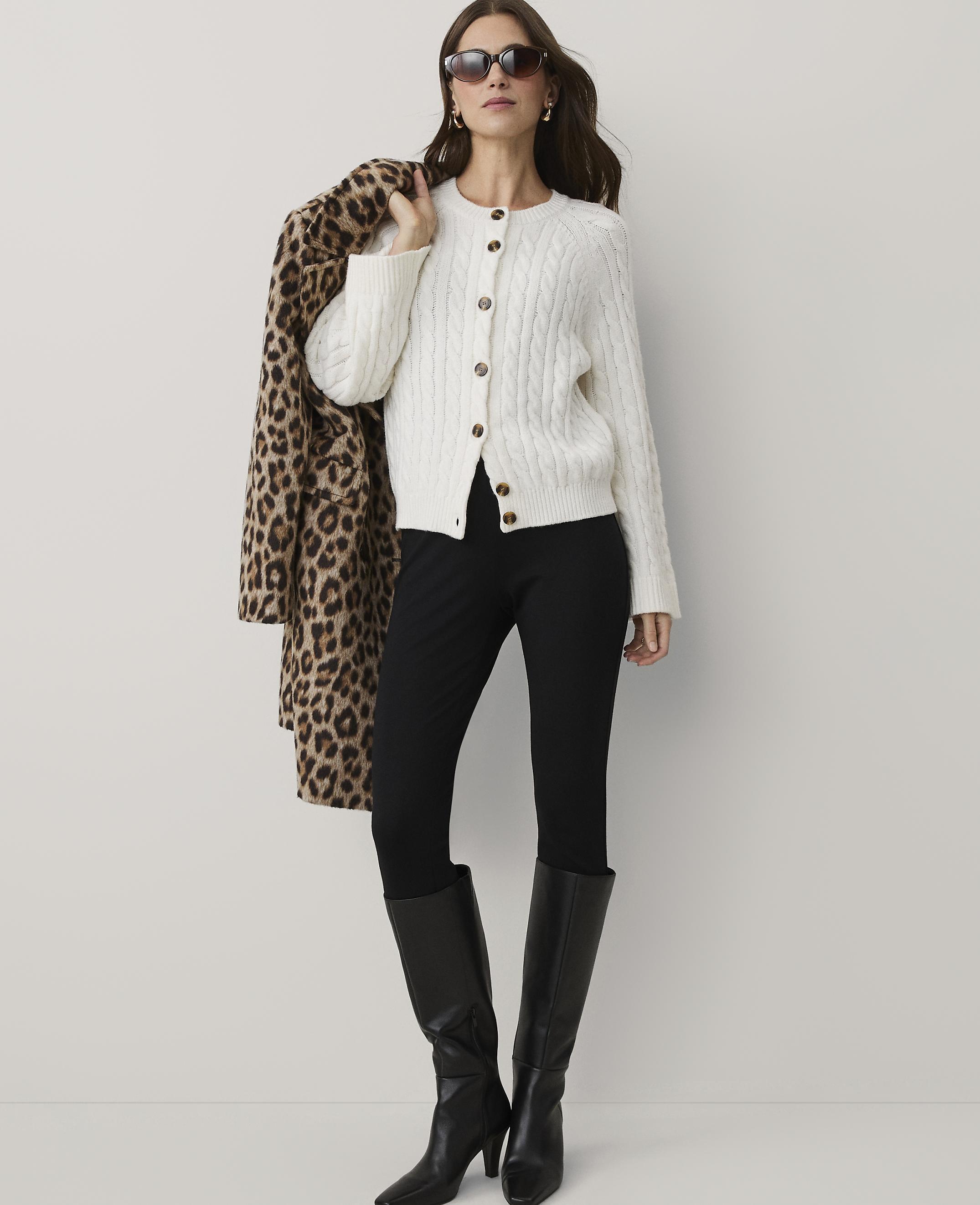 A woman wearing a white cable-knit cardigan, black pants, and black boots stands against a plain white background while holding a leopard-print coat over her shoulder.