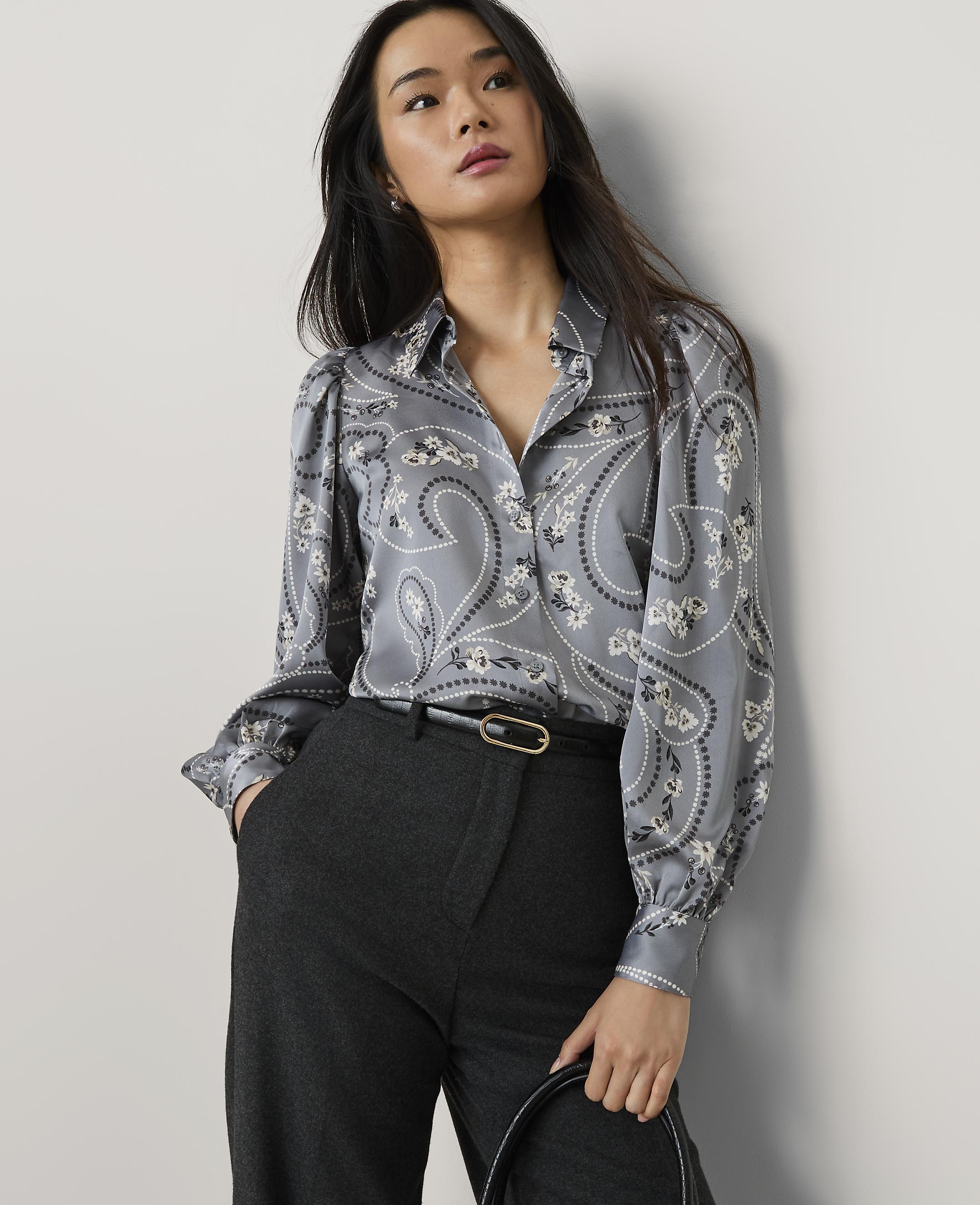 A woman wearing a grey paisley print blouse and dark trousers stands against a plain light grey wall.