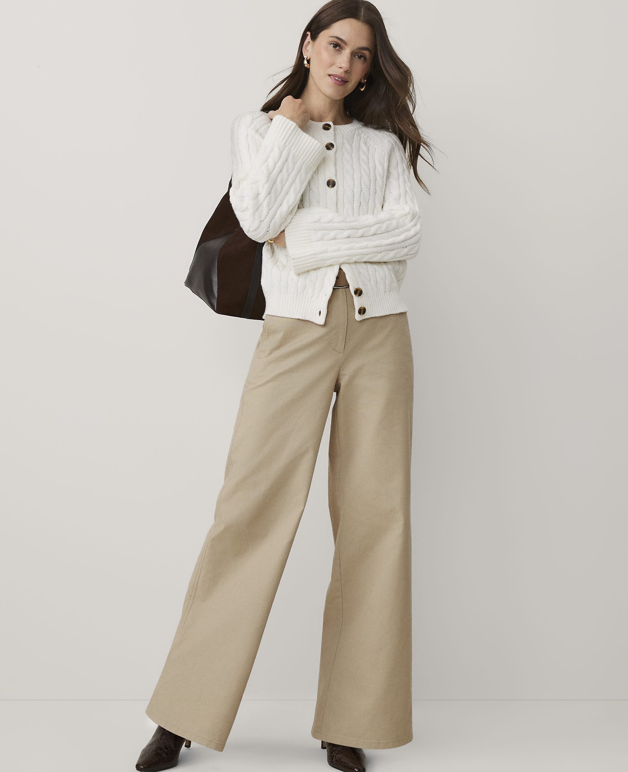 A woman wearing a white cable-knit cardigan and wide-leg khaki trousers stands against a plain white background.