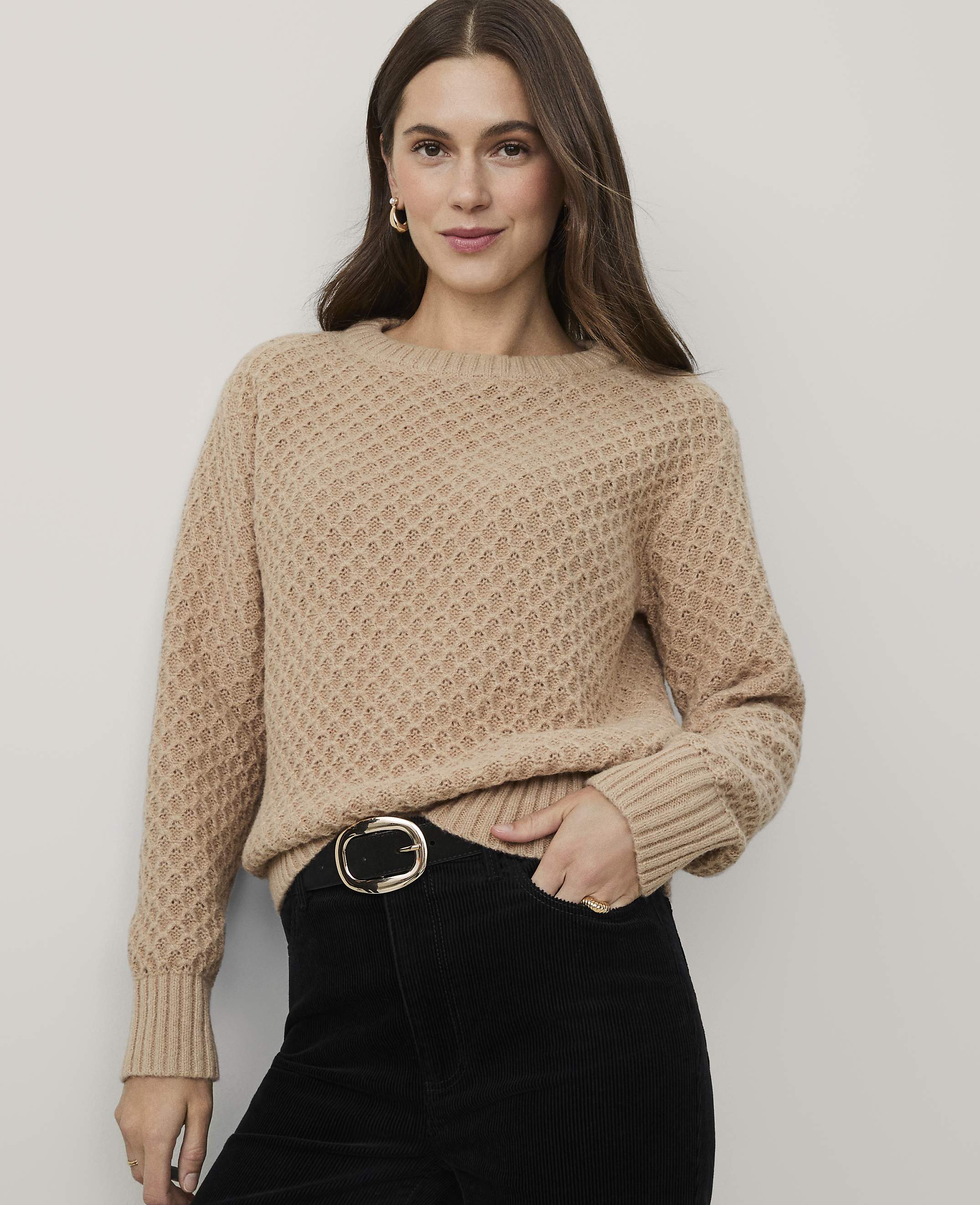 A woman wearing a tan textured sweater and black corduroy pants stands against a plain light gray background.
