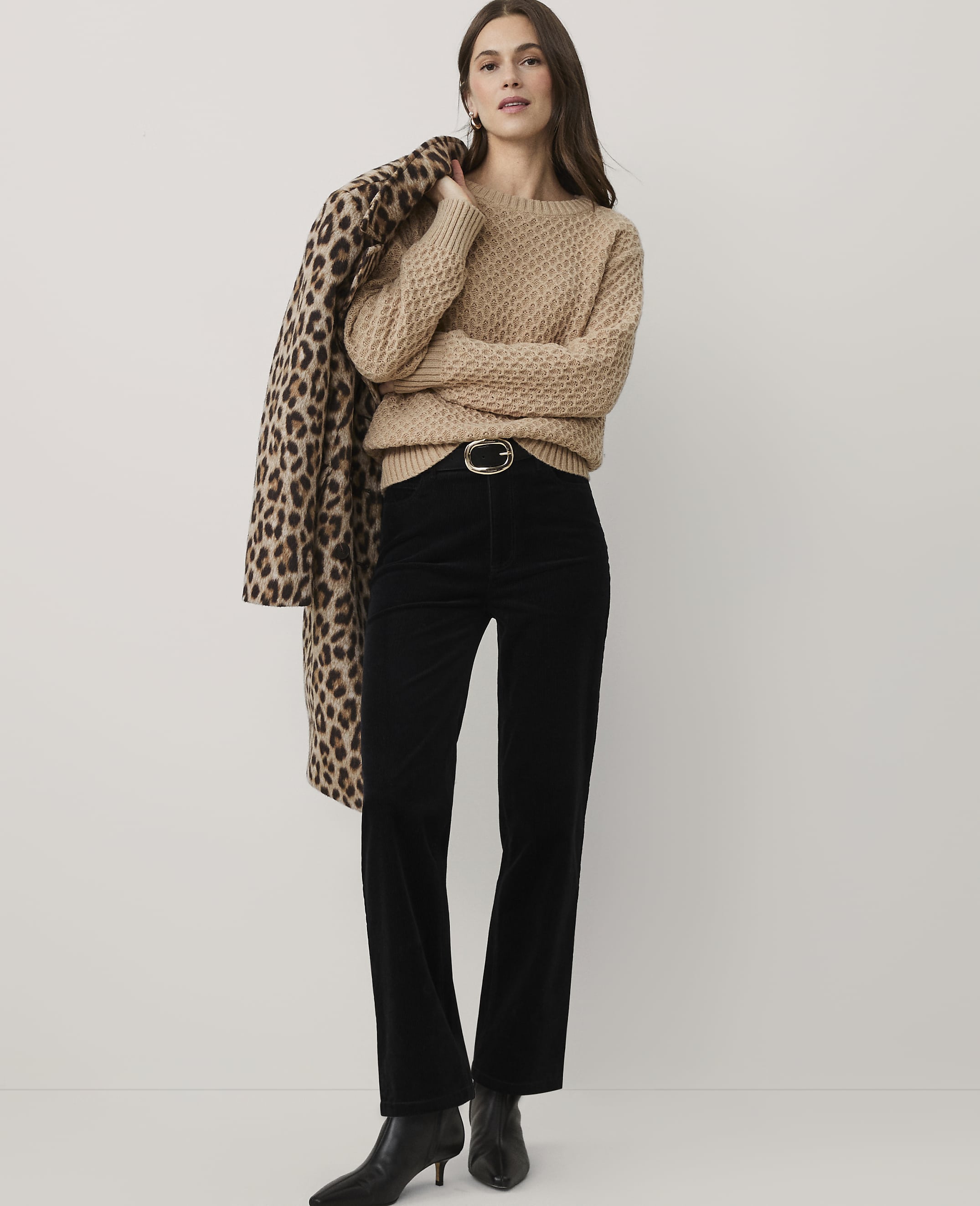 A woman wearing a tan sweater, black trousers, and black boots stands against a plain white background while draping a leopard-print coat over her shoulder.