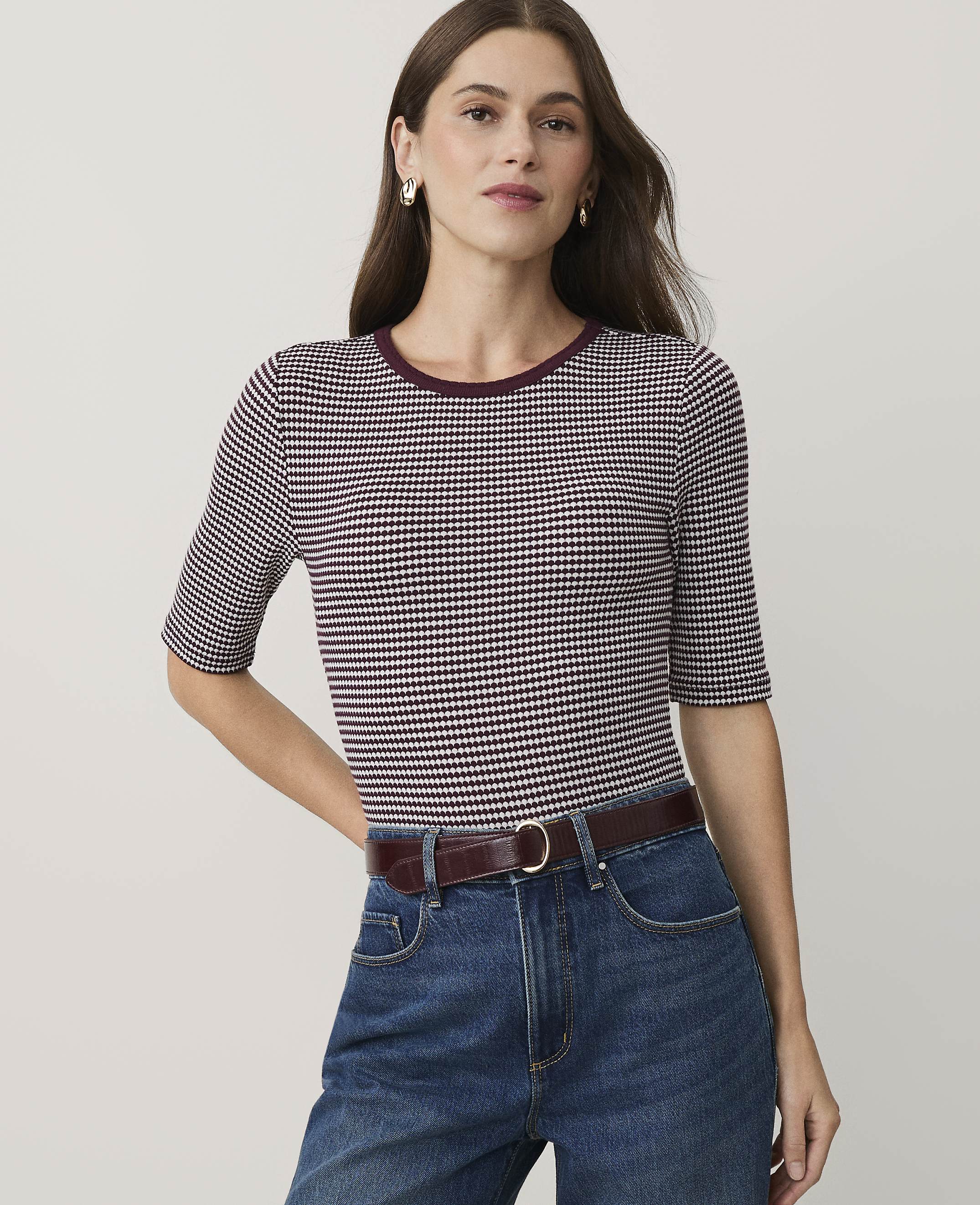 A woman wearing a purple and white patterned top and blue jeans stands against a plain, light-colored background.