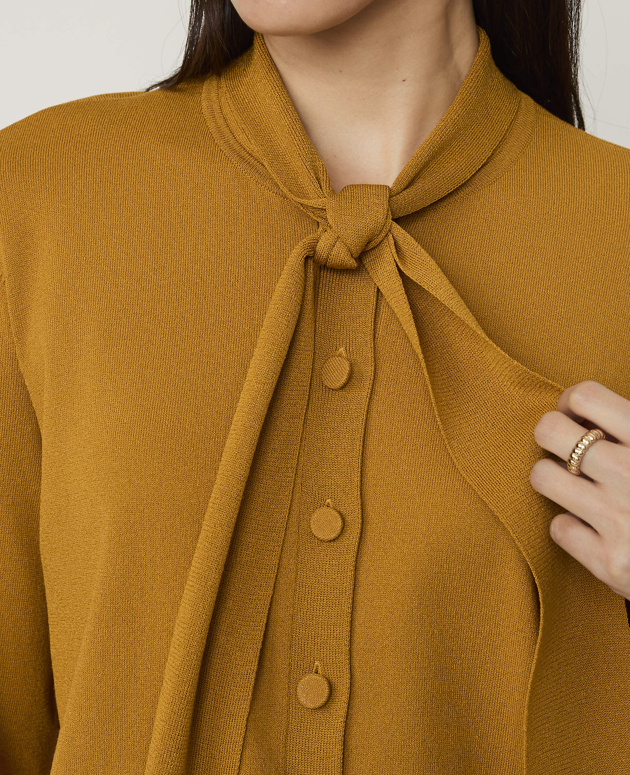 A person wears a mustard-yellow knit cardigan with a tie-neck detail against a plain white background.