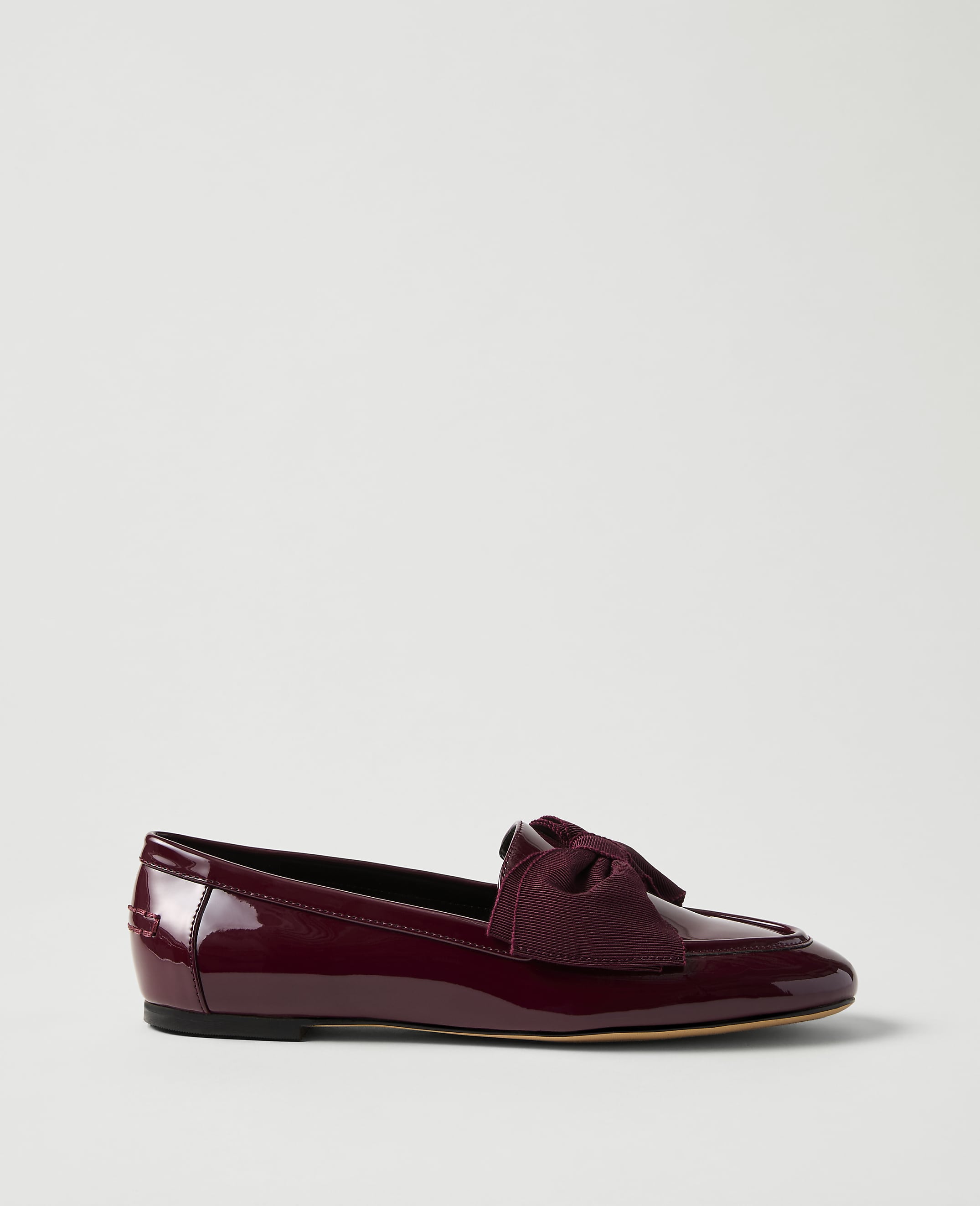 A burgundy patent leather flat shoe with a bow sits in the foreground against a plain, light gray background.