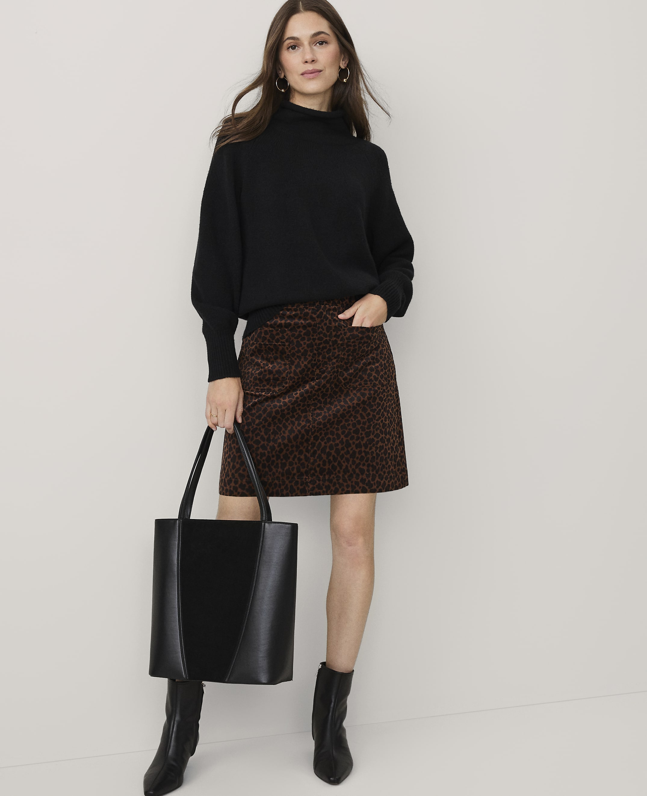 A woman wearing a black sweater, leopard-print skirt, and black boots stands holding a black tote bag against a plain white background.