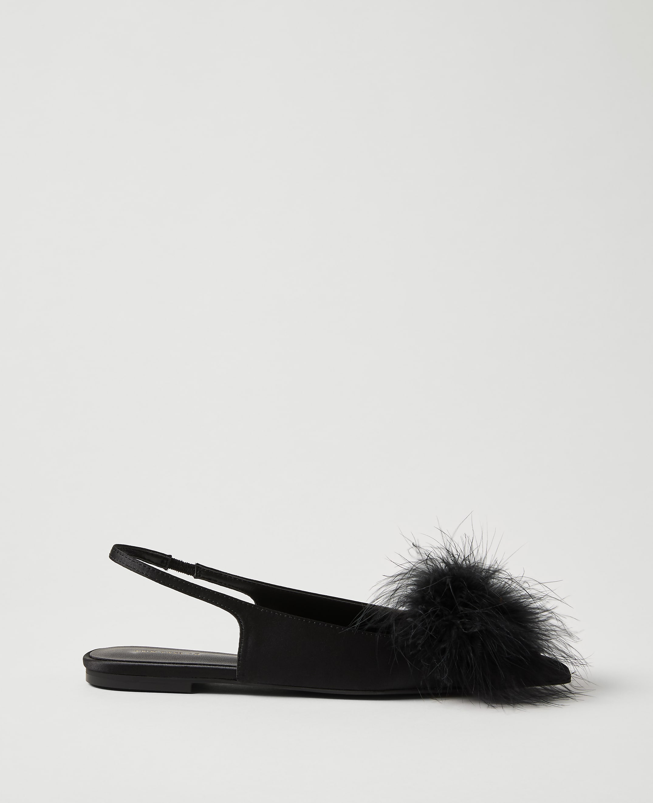 A black flat shoe with a fluffy pom-pom sits in the foreground against a plain, light gray background.