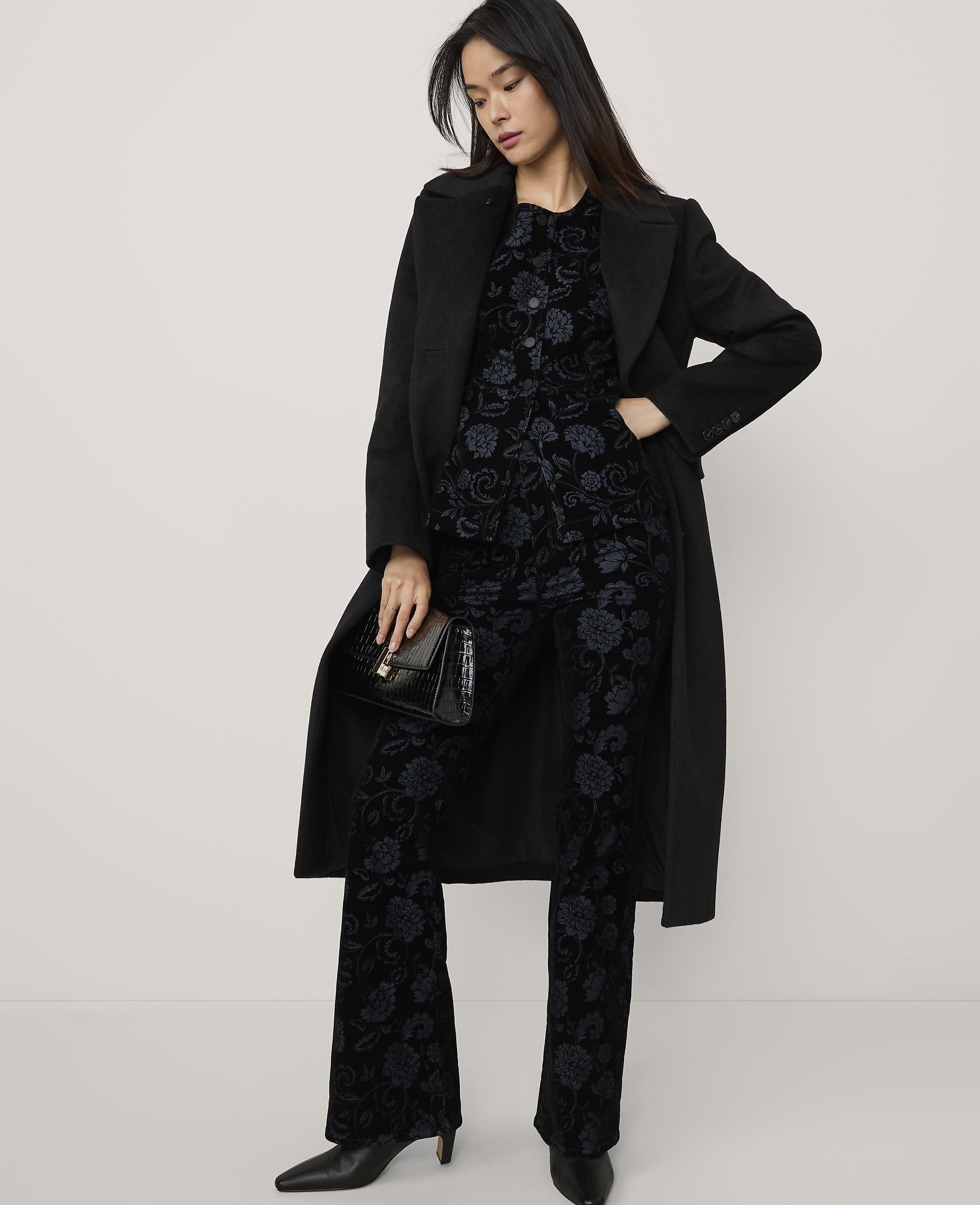 A woman wearing a black coat over a dark floral patterned suit and holding a clutch stands against a plain, light-colored background.