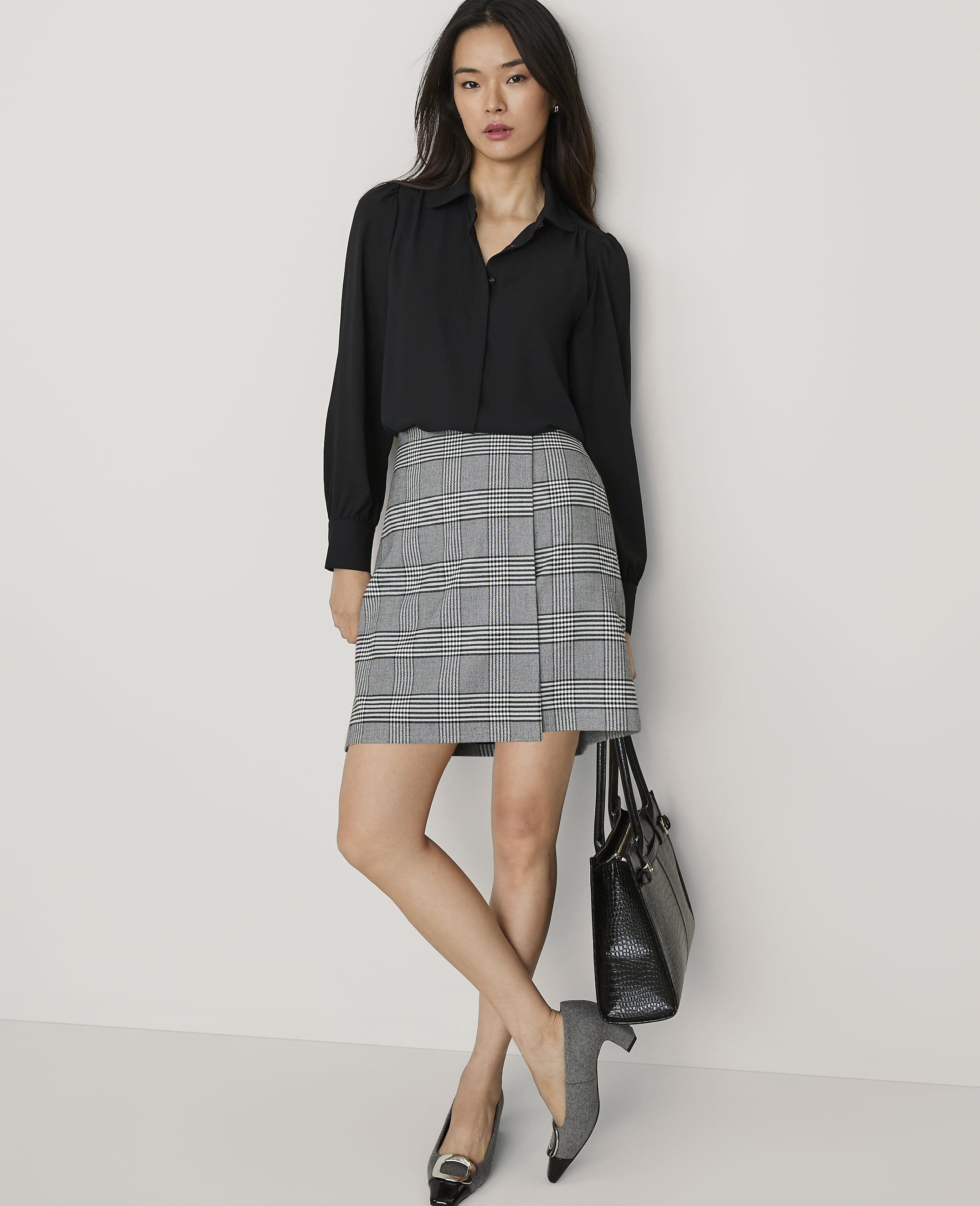 A woman wearing a black blouse and a grey plaid skirt stands against a plain white wall while holding a black handbag.