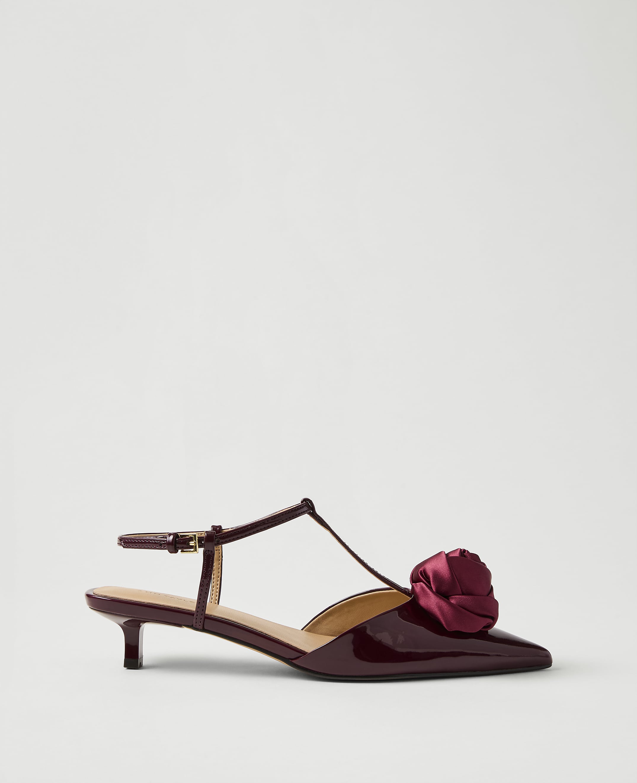 A burgundy patent leather kitten heel with a fabric rose detail sits against a plain, light grey background.