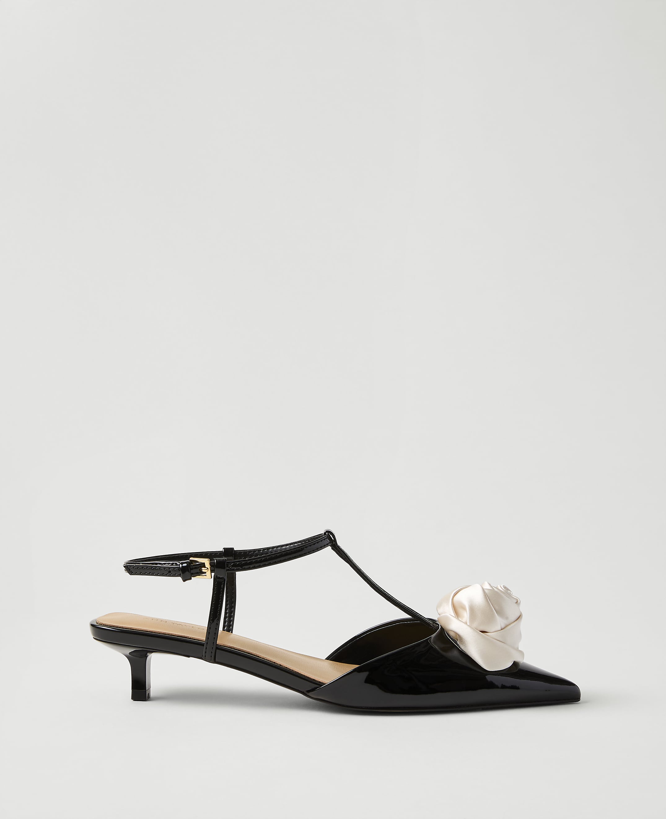 A black patent leather kitten heel with a cream-colored fabric flower on the toe sits against a plain, light gray background.