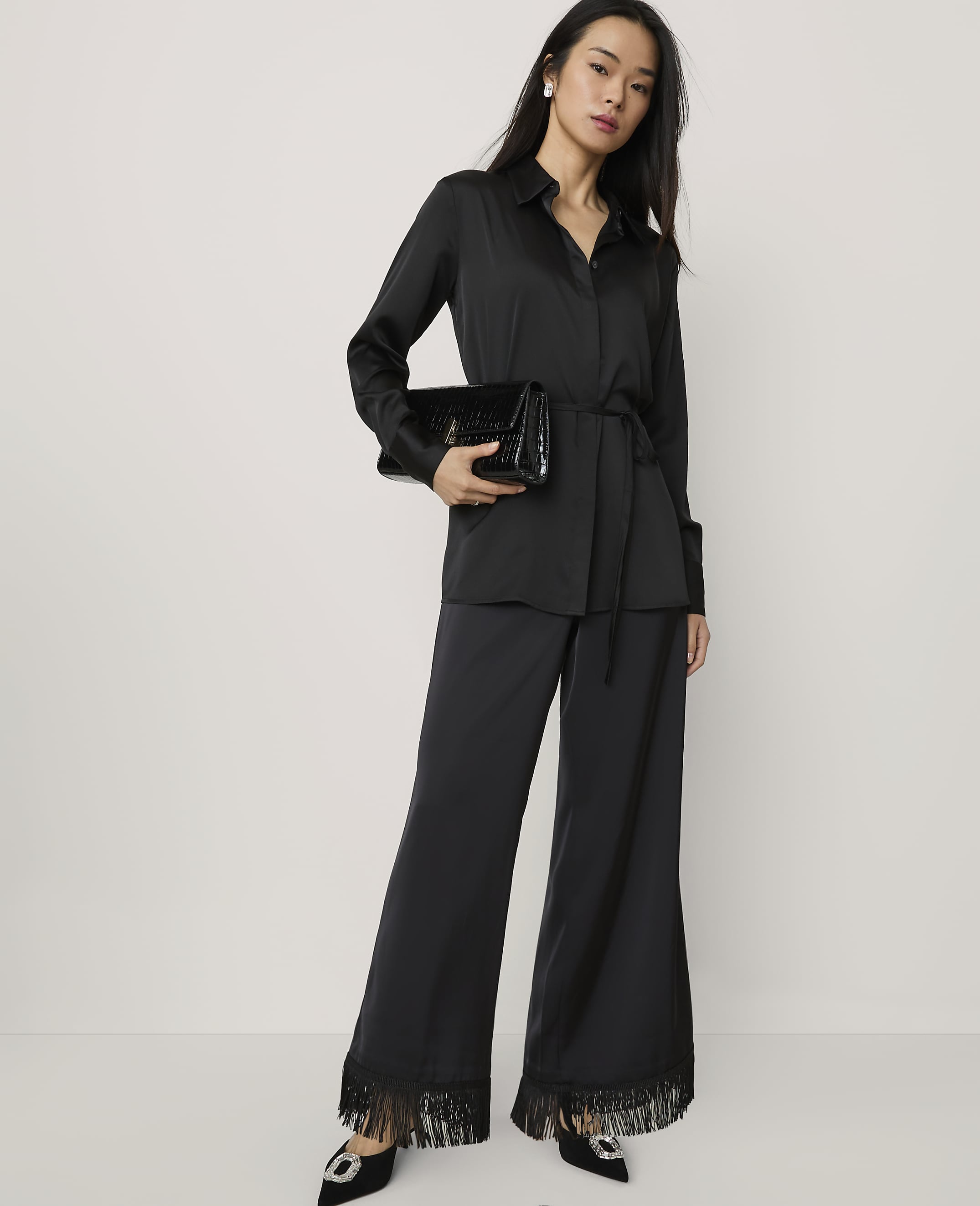 A woman wearing a black satin shirt and wide-leg fringed trousers stands against a plain white background.
