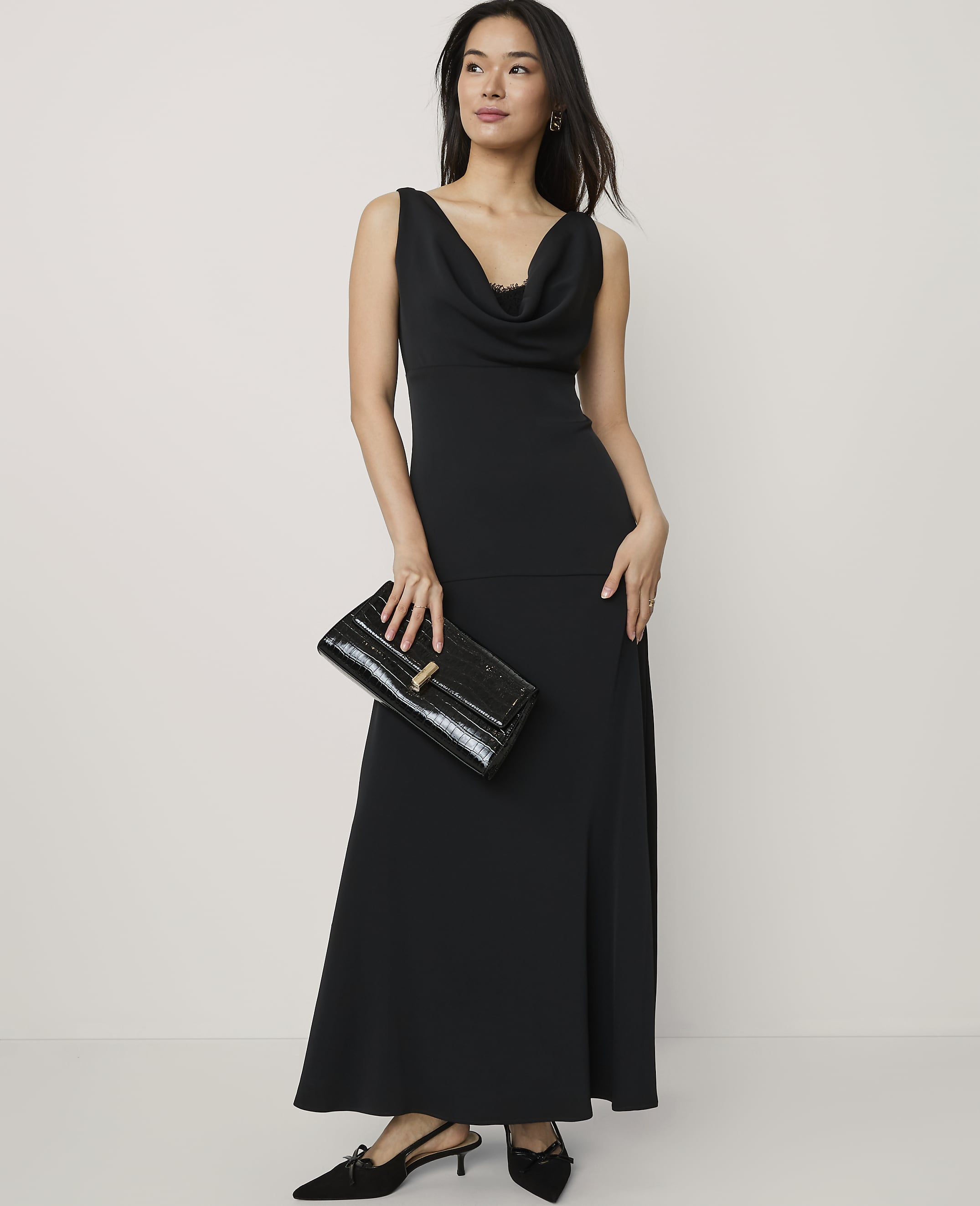 A woman wearing a long black cowl-neck dress and holding a black clutch stands against a plain, light-colored background.