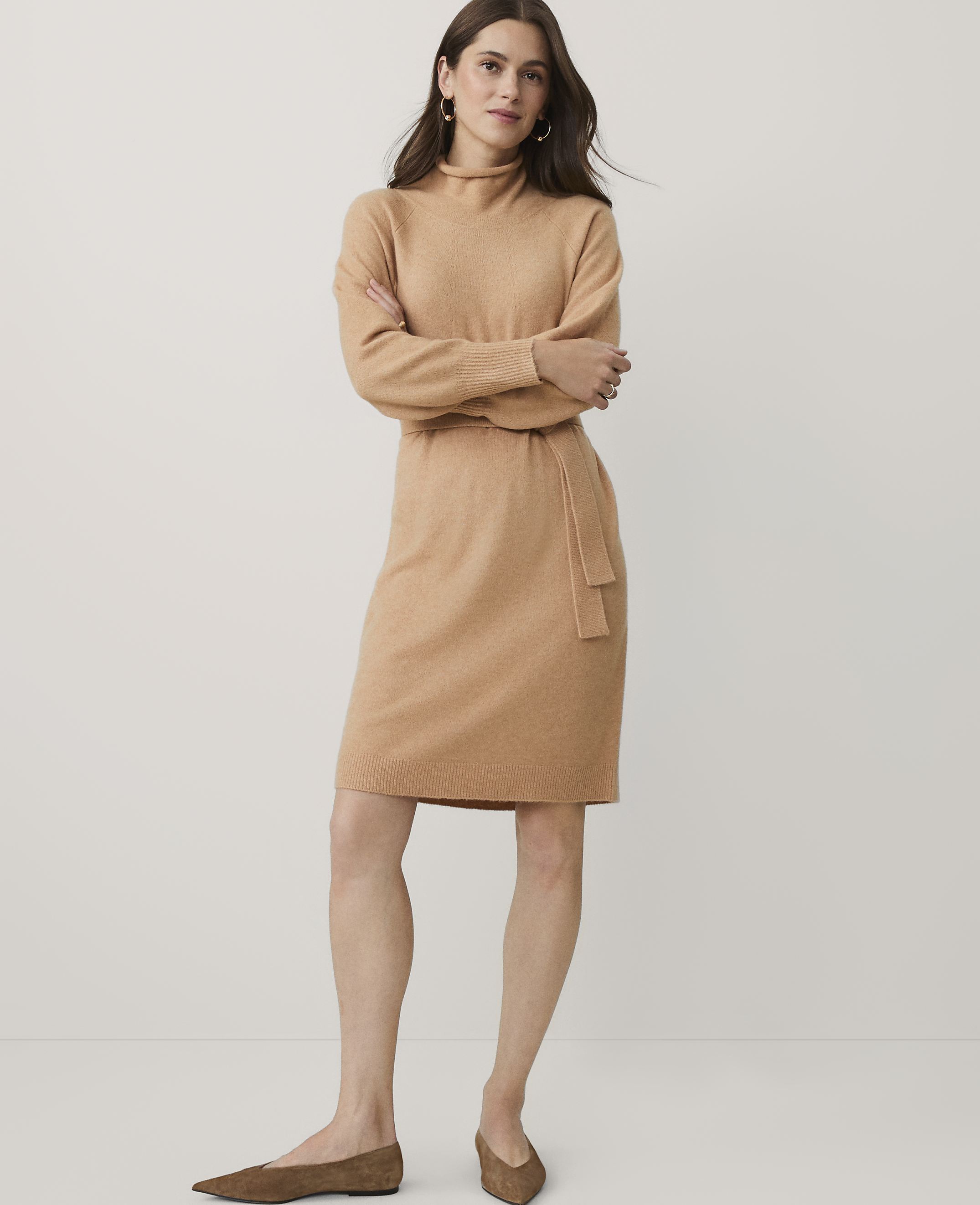 A woman wearing a tan knit dress and brown flats stands against a plain, light-colored background.
