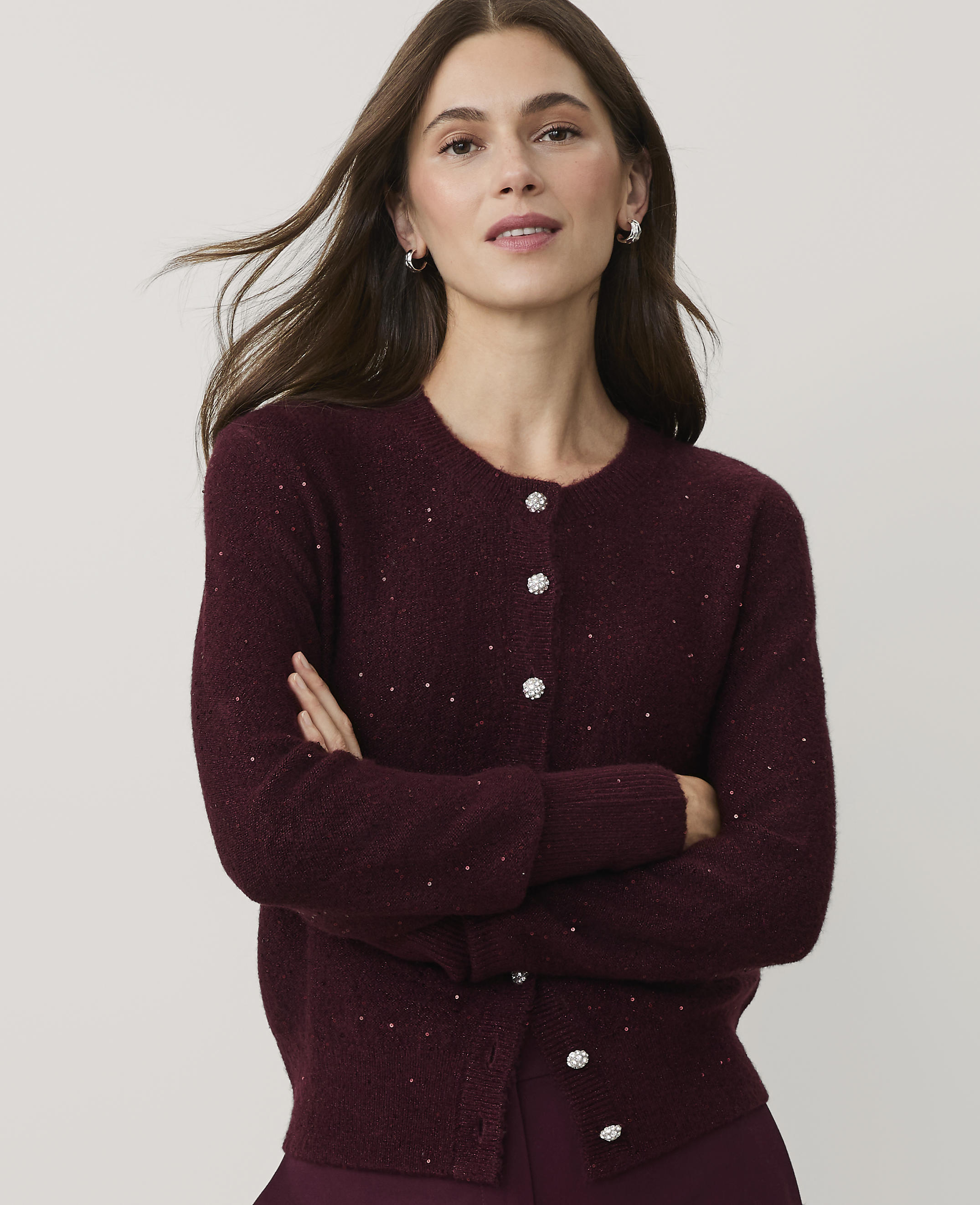 A woman with long brown hair wearing a sparkling burgundy cardigan stands with her arms crossed against a plain light gray background.