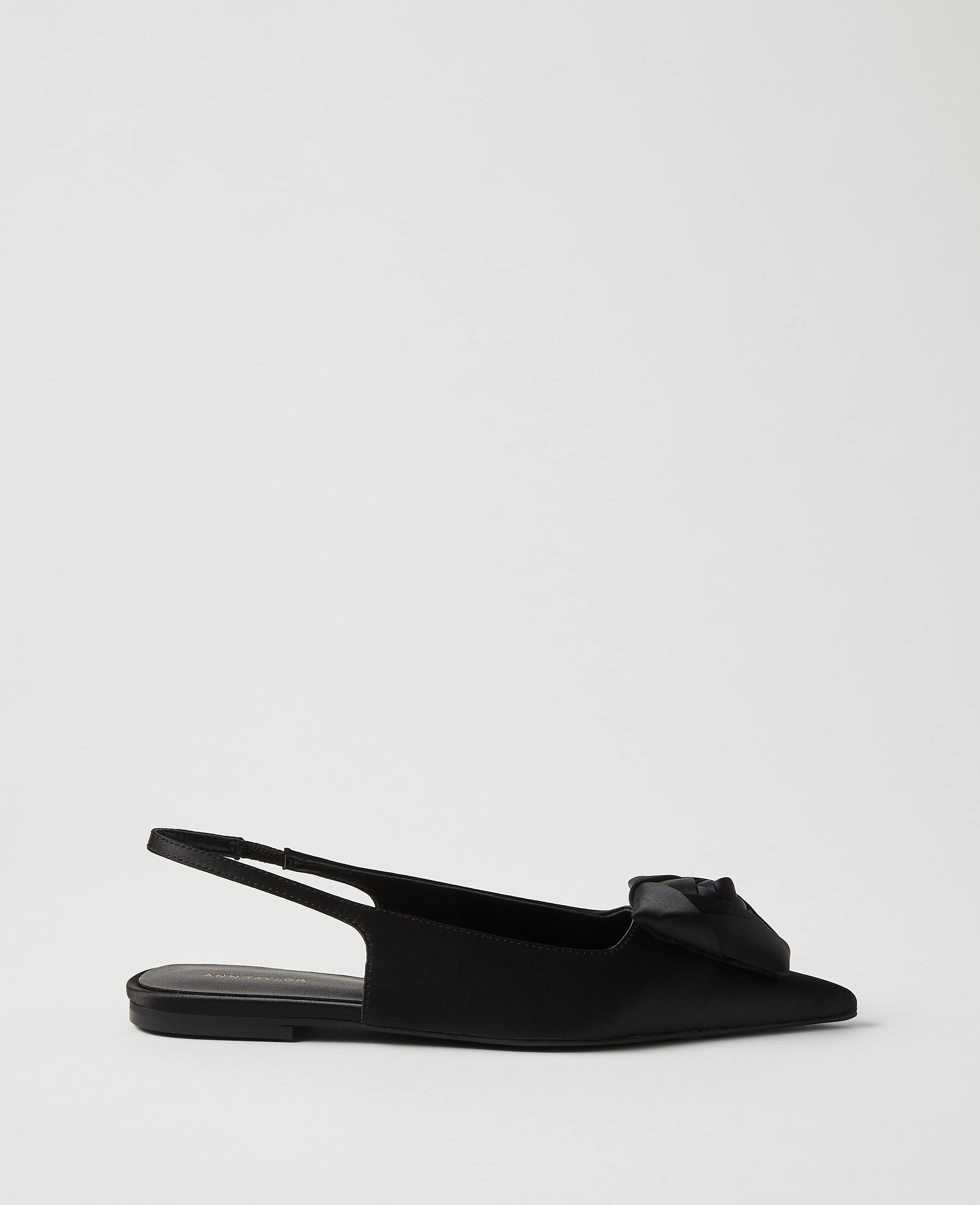 A single black satin slingback flat with a bow on the toe sits against a plain, light gray background.