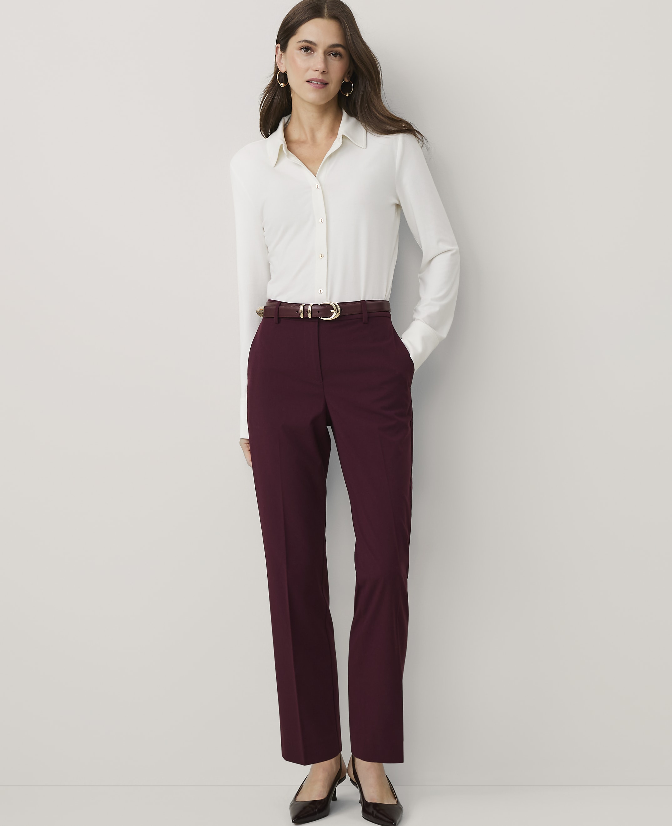 A woman wearing a white button-down shirt and burgundy trousers stands against a plain white background.