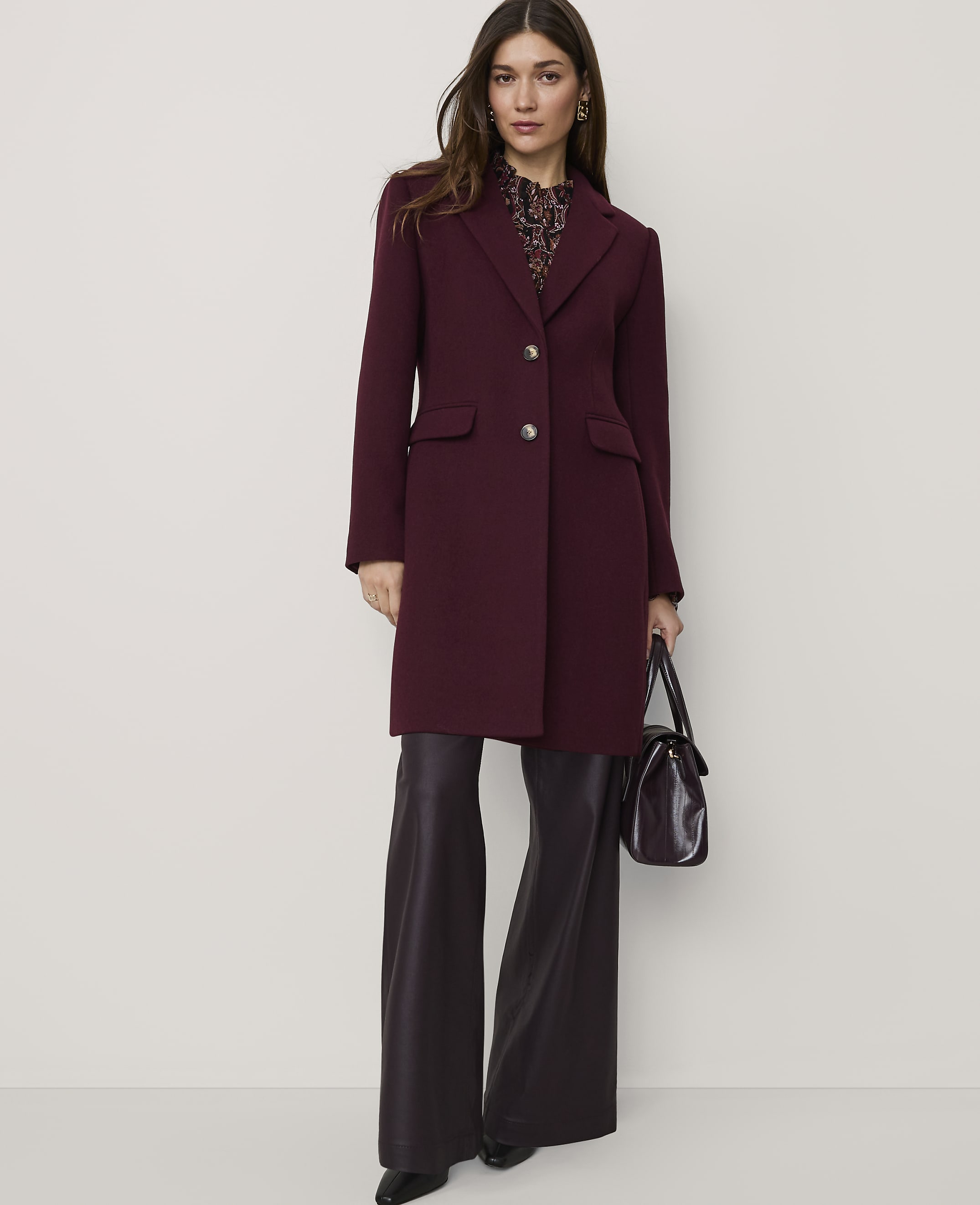 A woman wearing a long burgundy coat and wide-leg dark purple trousers stands against a plain, light-colored background.