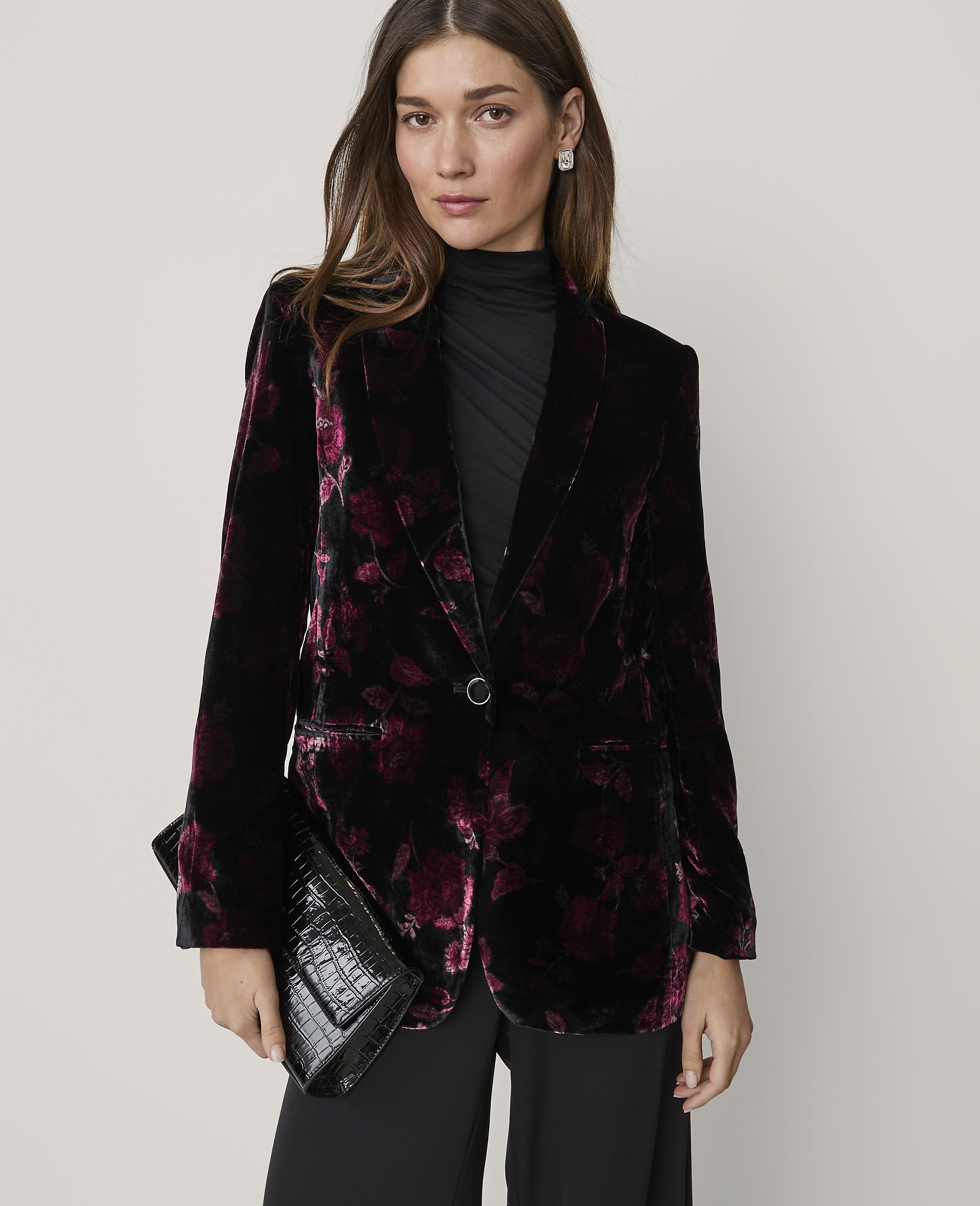 A woman wearing a floral velvet blazer and black clothing holds a black clutch against a plain, light-colored background.