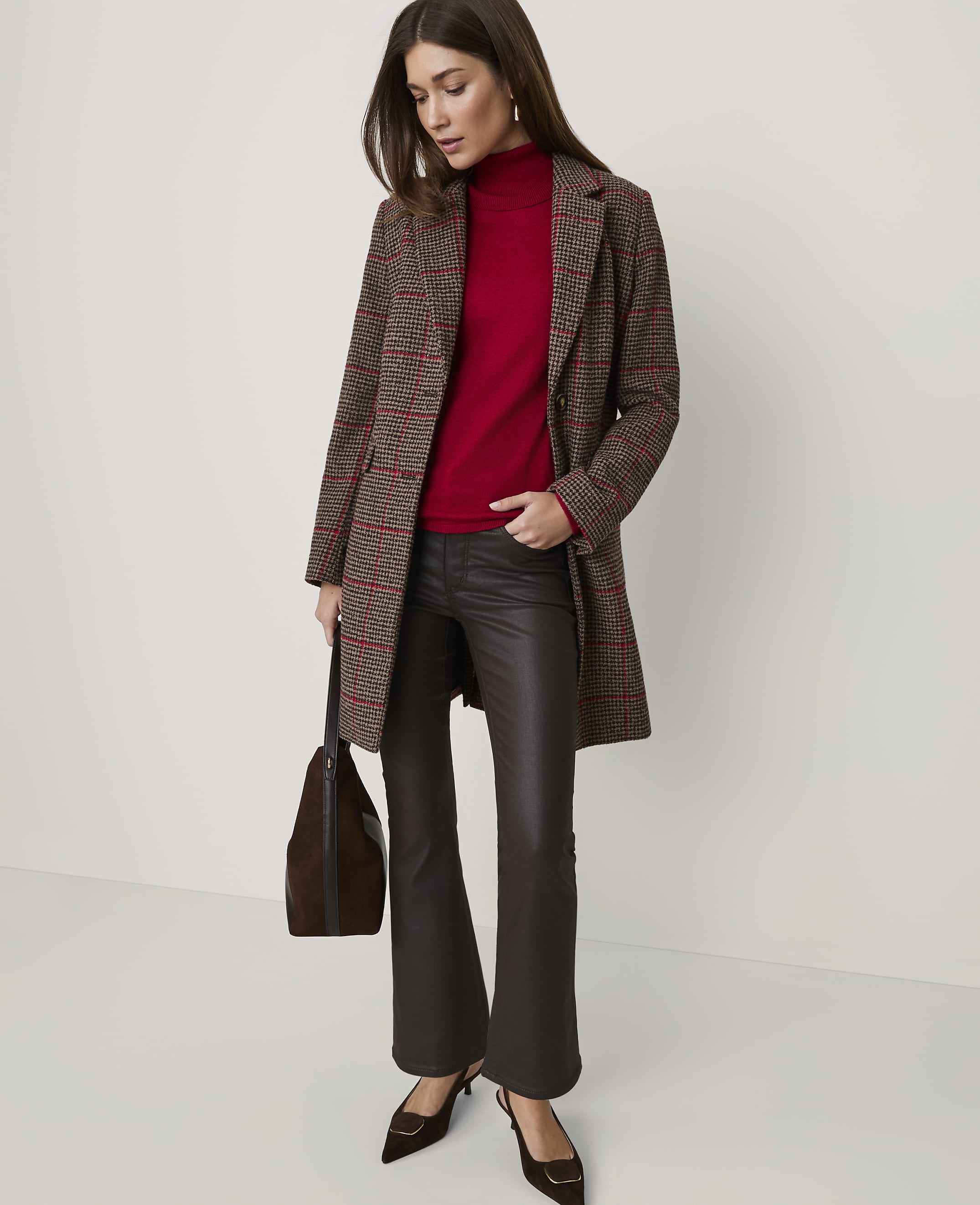 A woman wearing a plaid coat, red turtleneck, and dark faux-leather trousers stands against a plain, light-colored background.