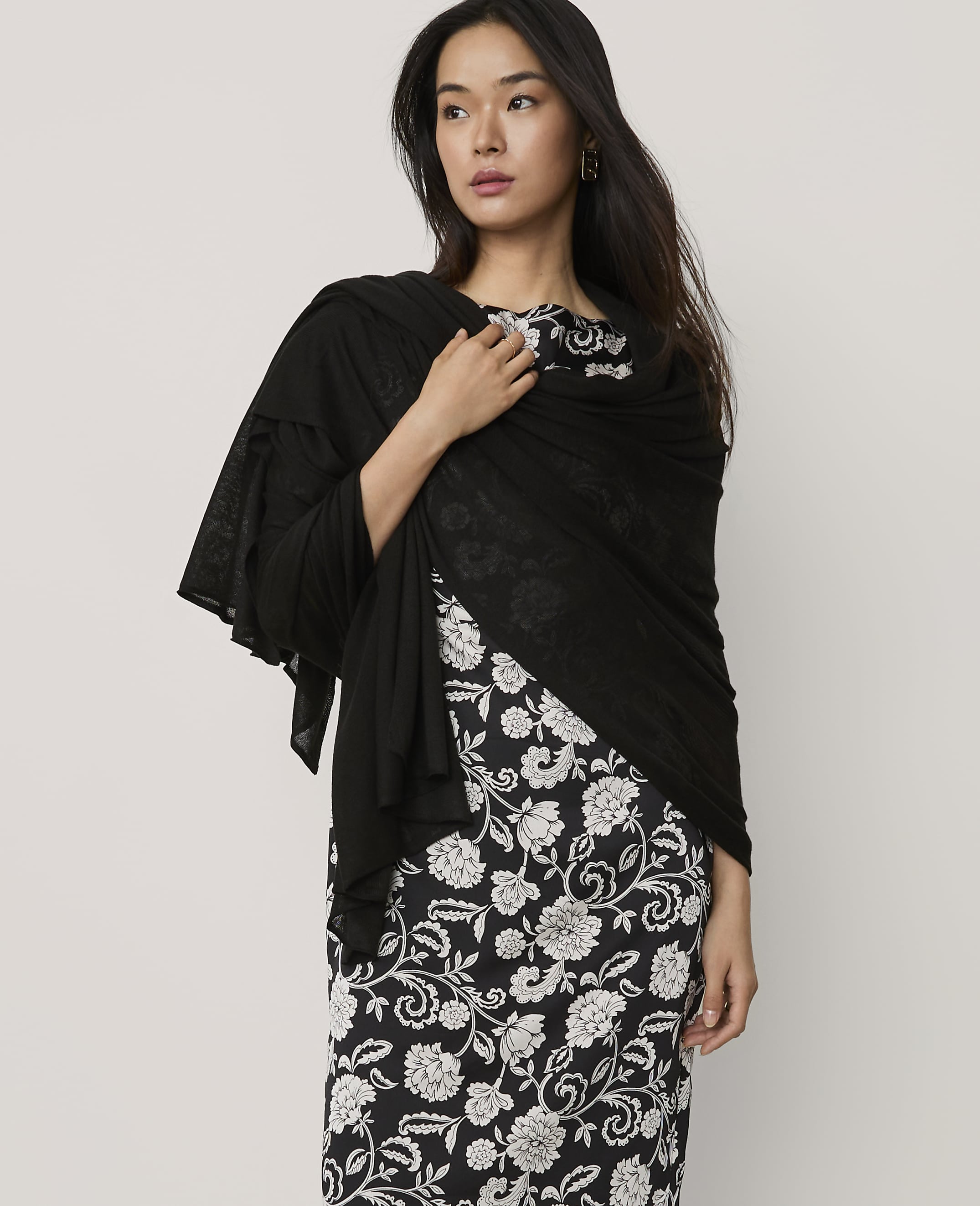 A woman wearing a black shawl over a black and white floral dress stands against a plain light gray background.