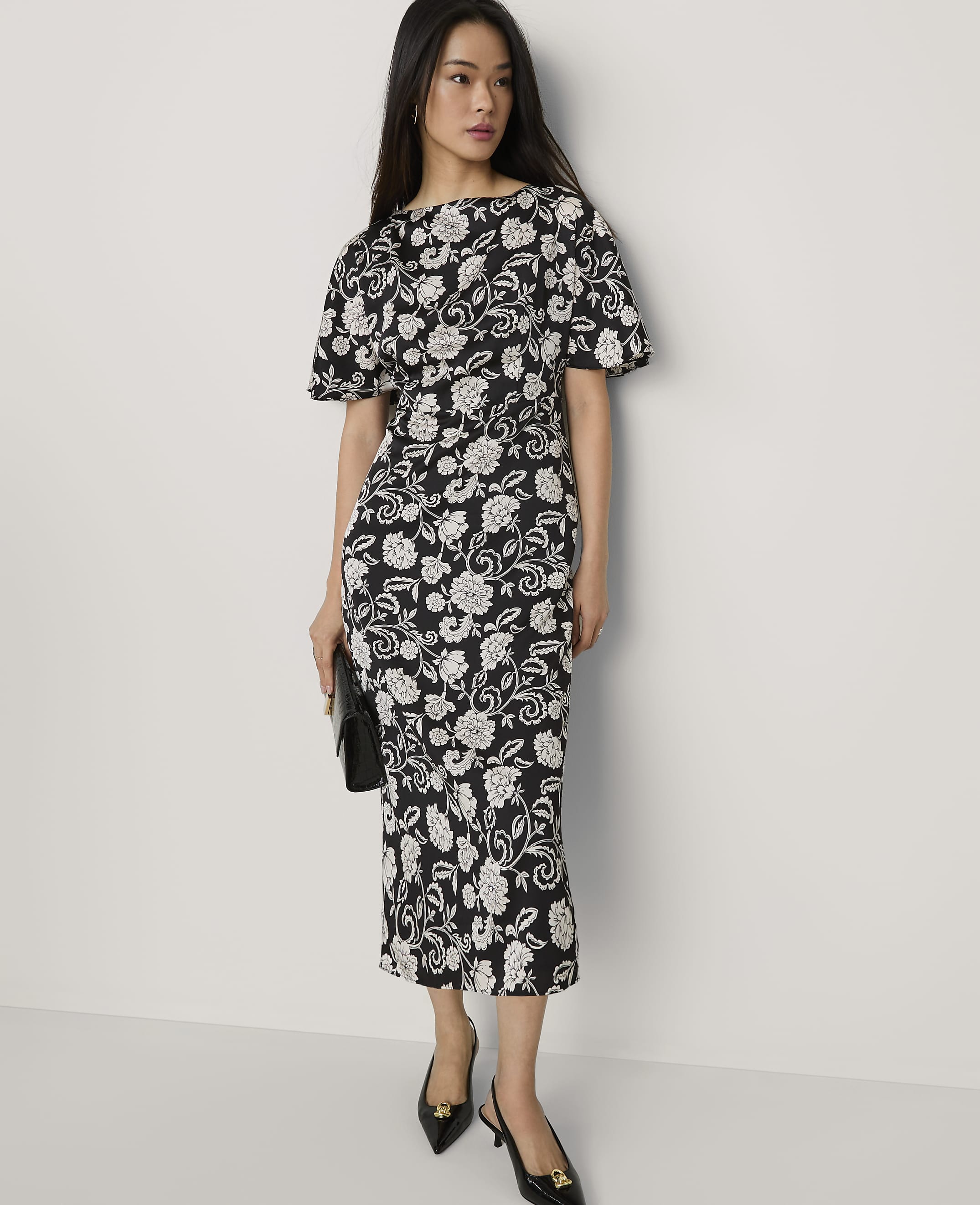 A woman wearing a black and white floral midi dress and black heels stands against a plain white background.