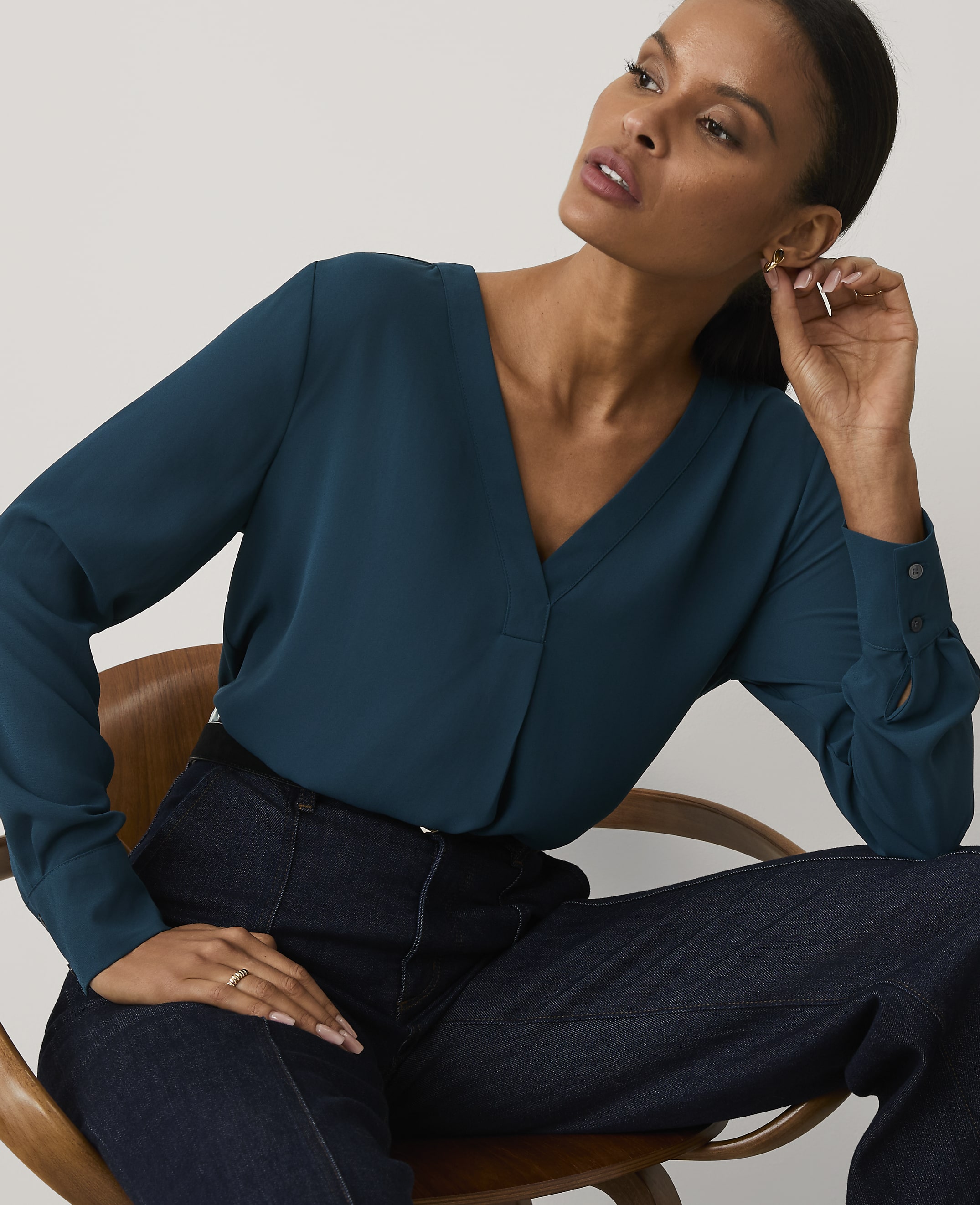 A woman wearing a teal V-neck blouse and dark blue jeans sits in a wooden chair against a plain, light-colored background.