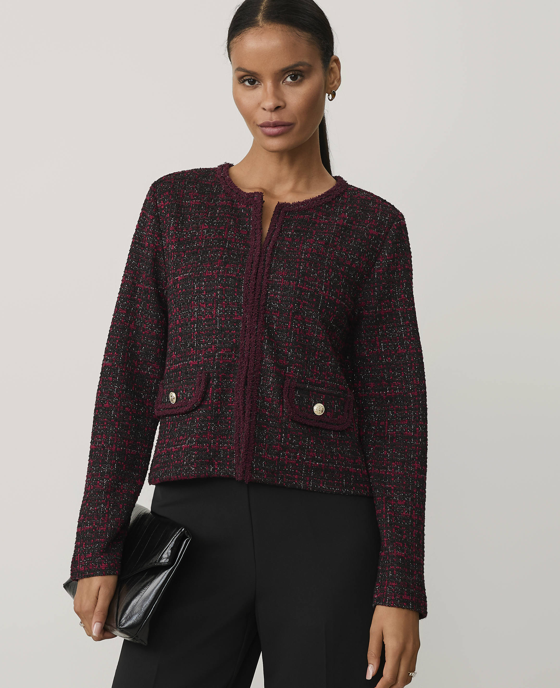 A woman wearing a burgundy tweed jacket and black trousers stands against a plain light gray background.