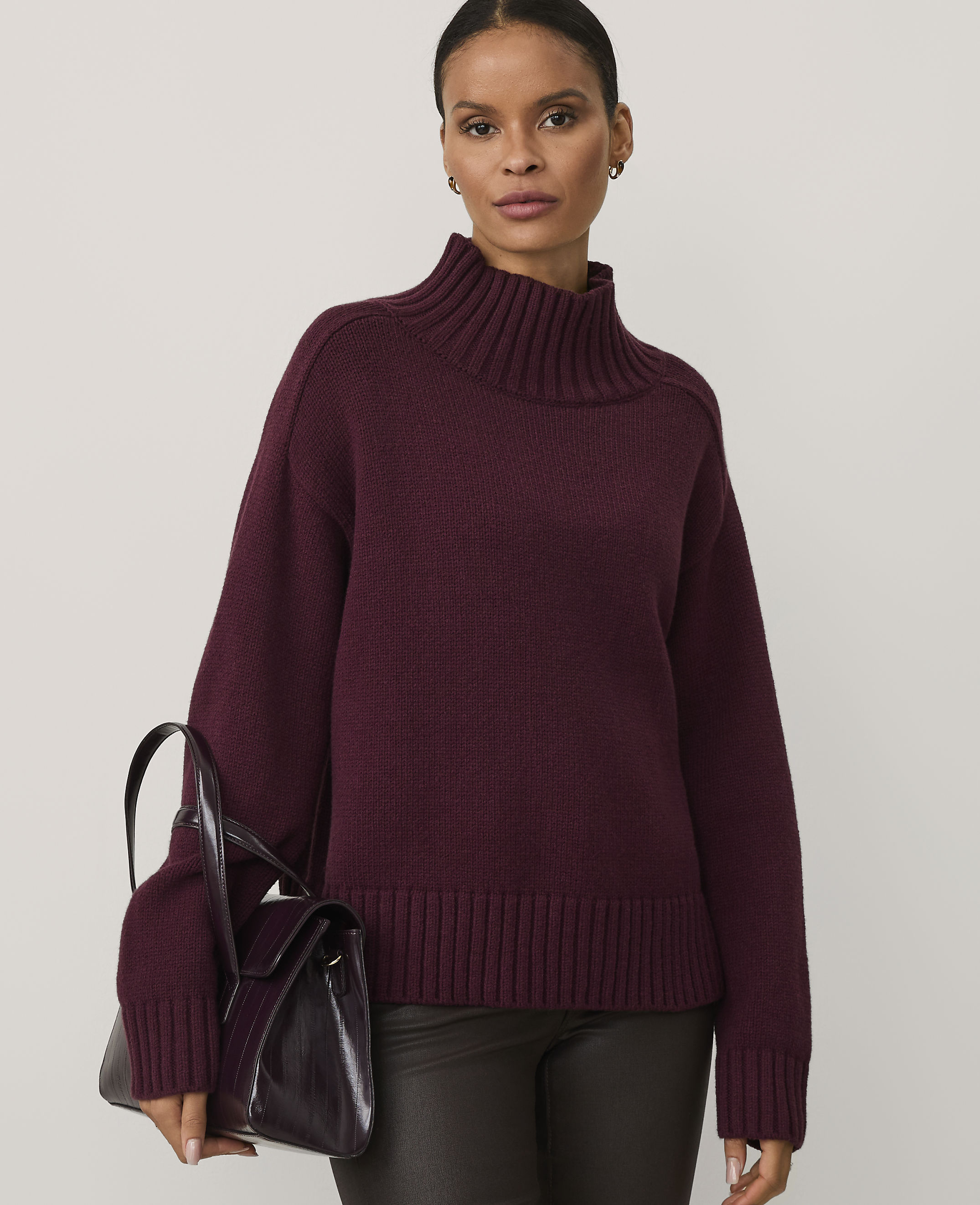 A woman wearing a burgundy turtleneck sweater and dark trousers holds a matching handbag against a plain light gray background.