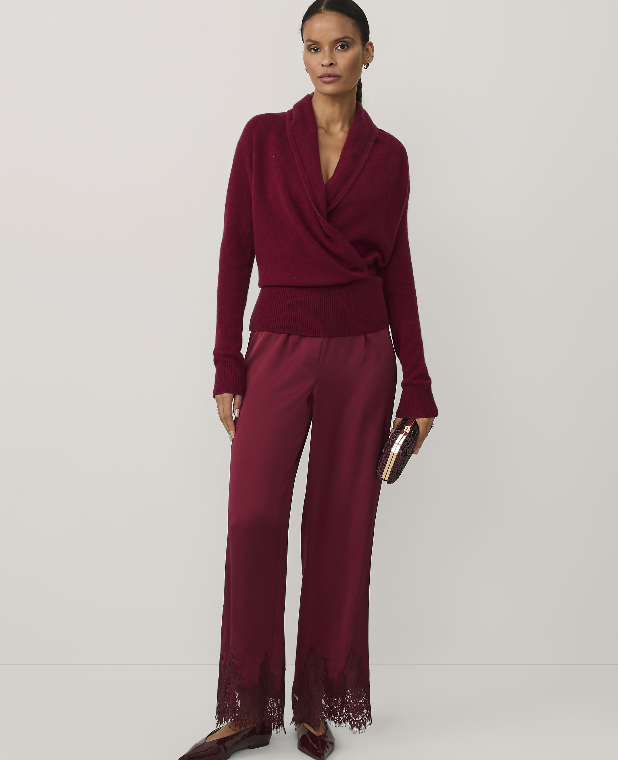 A woman wearing a burgundy wrap sweater and matching satin trousers with lace hems stands against a plain white background.