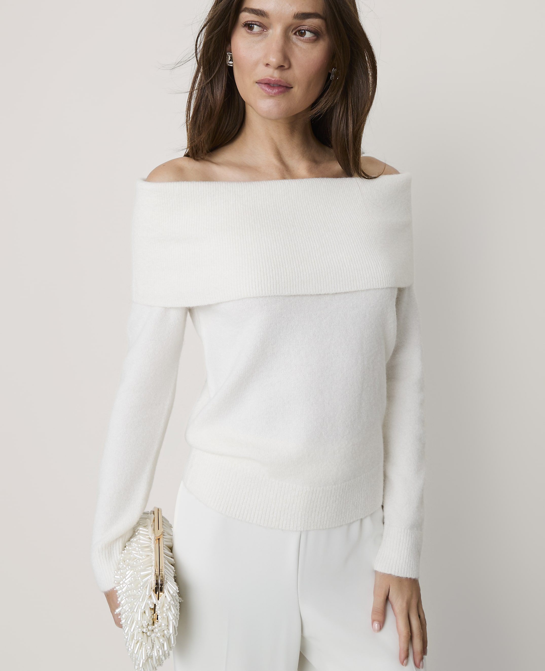 A woman wearing an off-the-shoulder white sweater and white trousers stands against a plain, light-colored background.