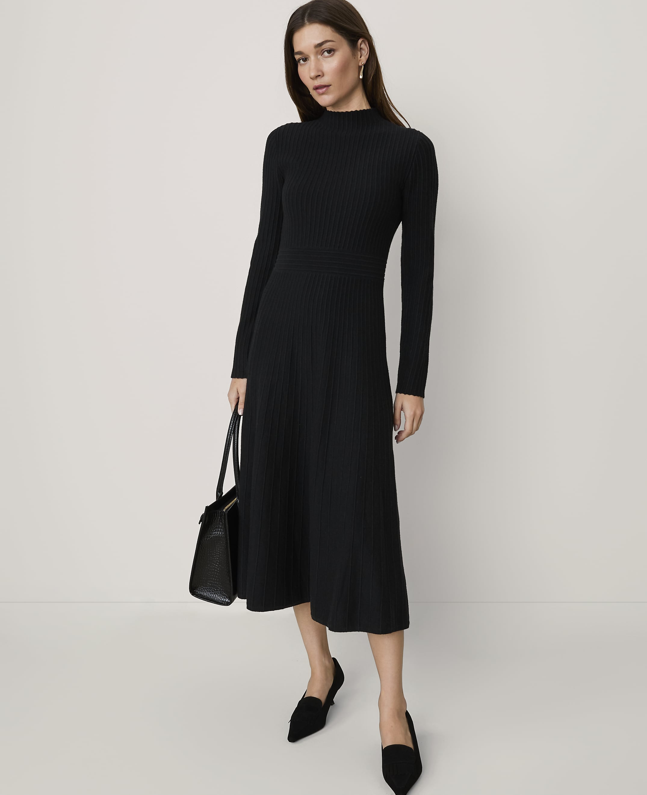 A woman wearing a long-sleeved black ribbed midi dress and black pointed-toe flats stands against a plain, light-colored background.