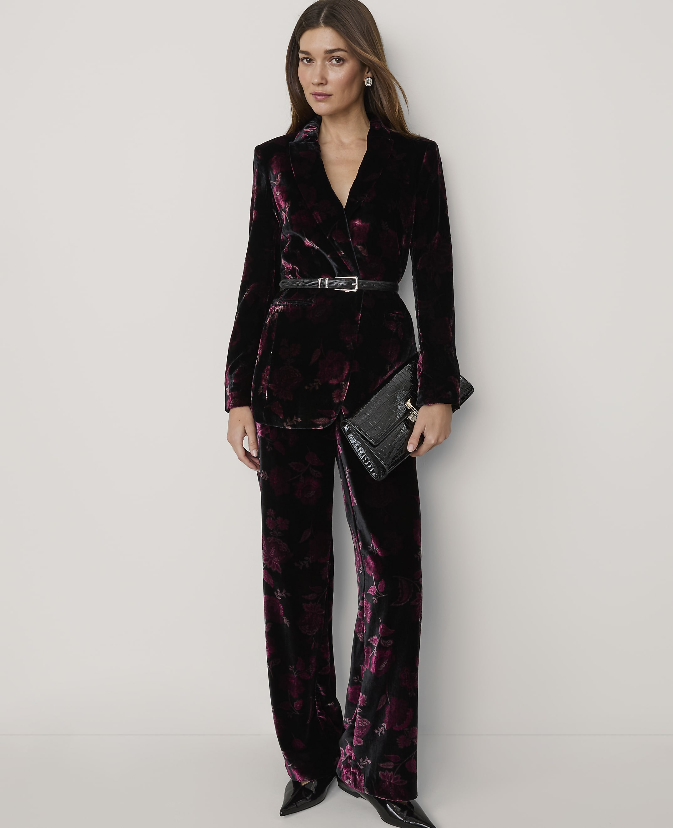 A woman wearing a dark purple velvet floral suit and holding a black clutch stands against a plain white background.