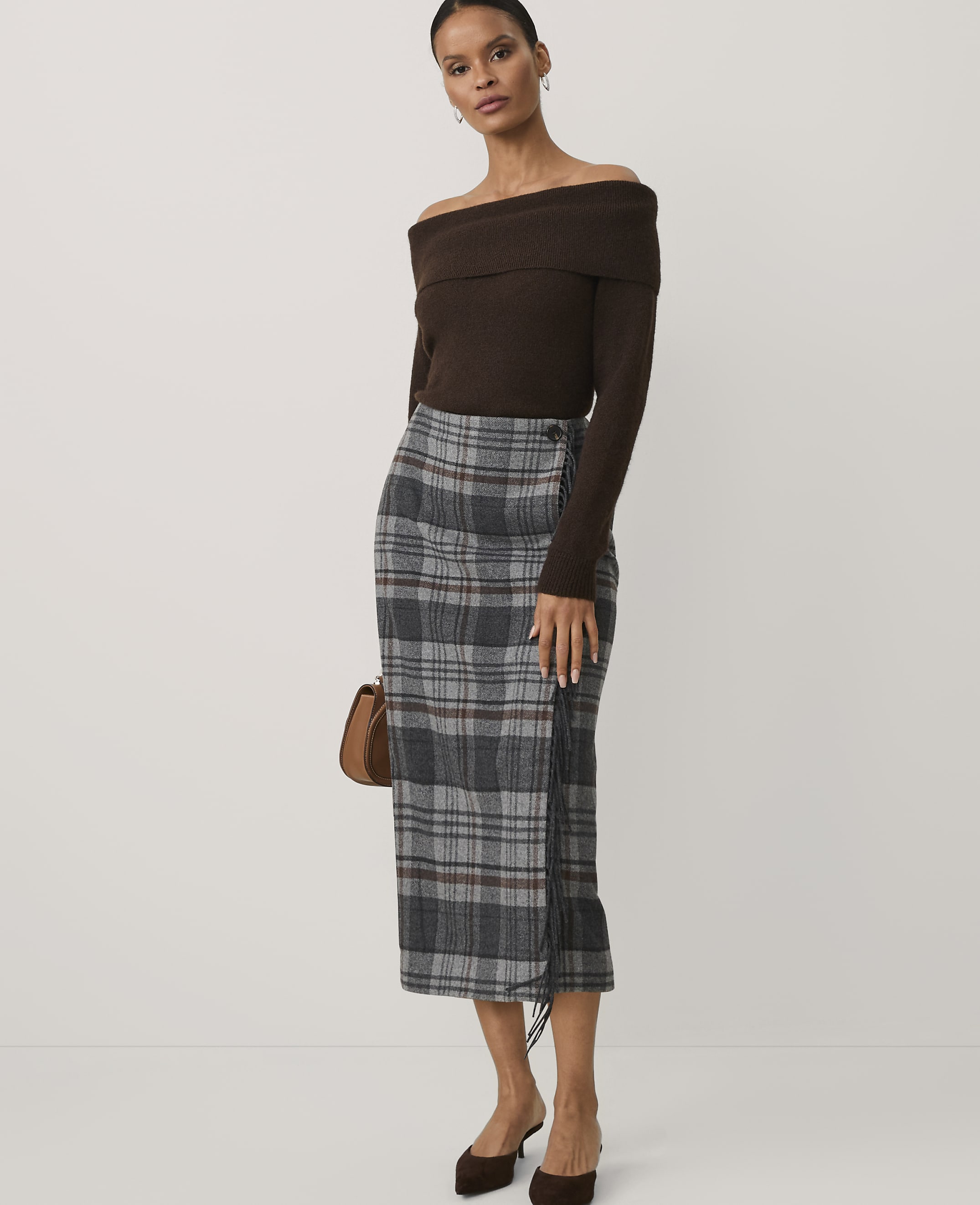A woman wearing an off-the-shoulder brown top and a grey plaid skirt stands against a plain white background.