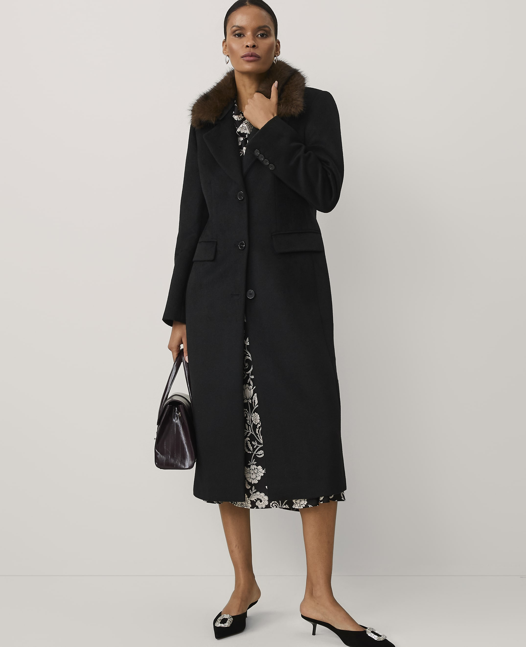 A woman wearing a long black coat with a faux-fur collar and a floral dress stands against a plain white background.