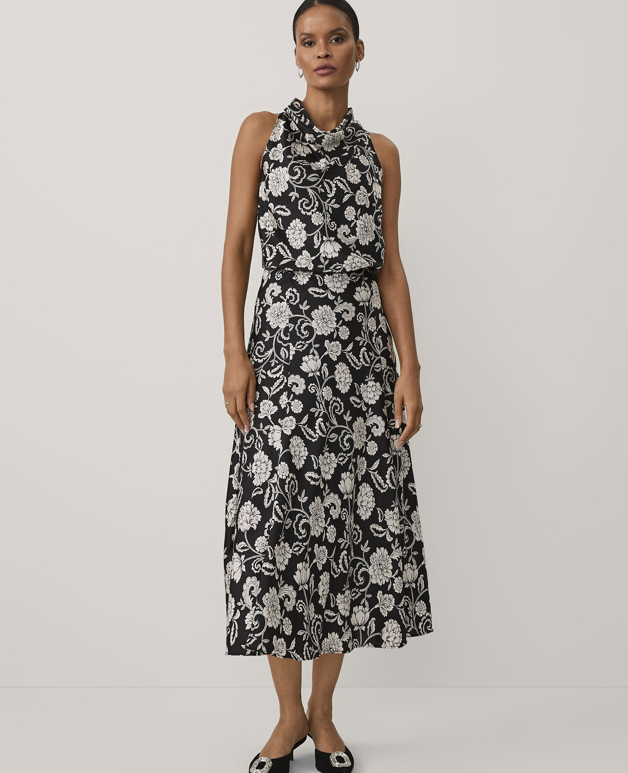 A woman wearing a black and white floral print dress and black heels stands against a plain white background.