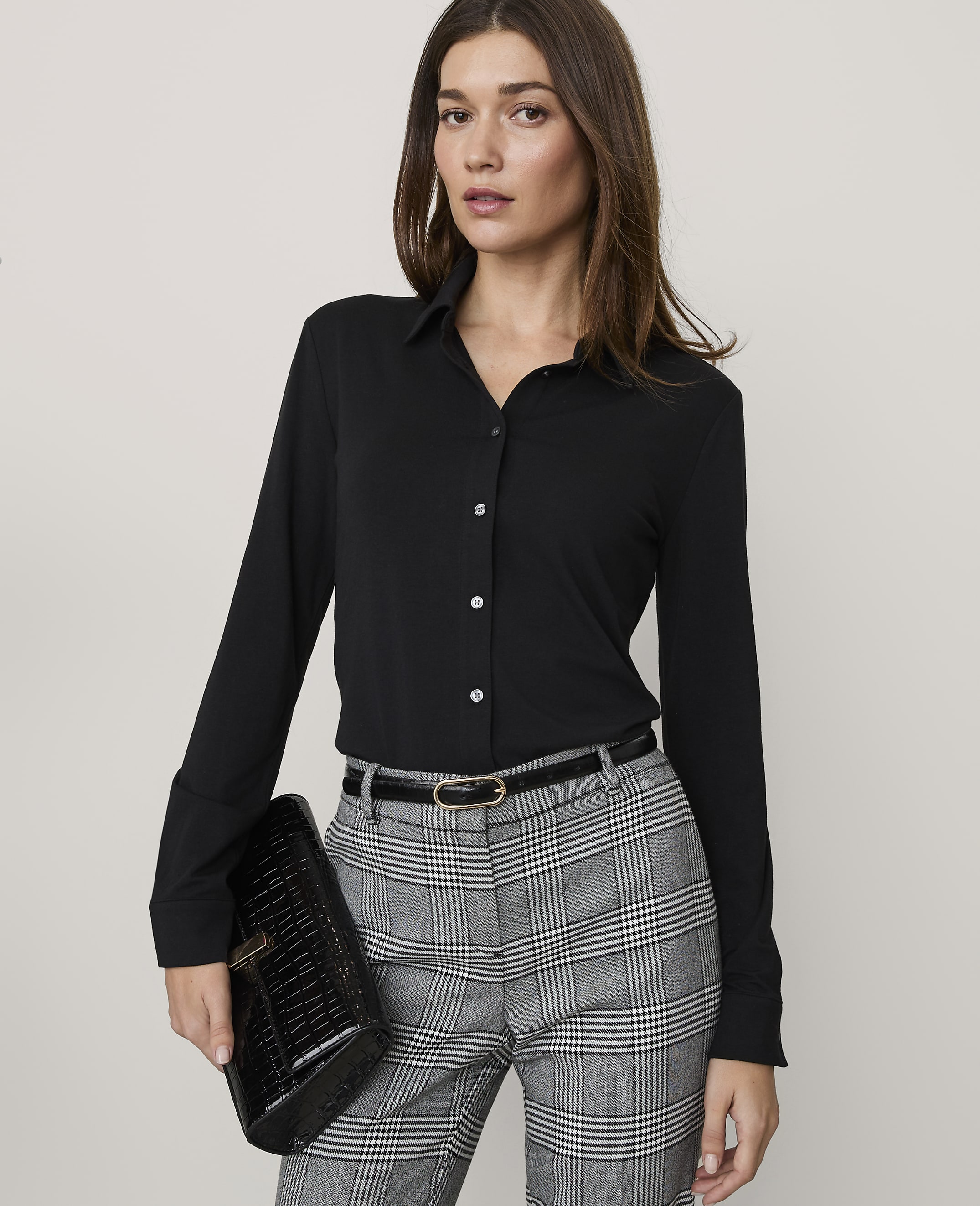 A woman wearing a black button-down shirt and plaid trousers stands against a plain, light-colored background.