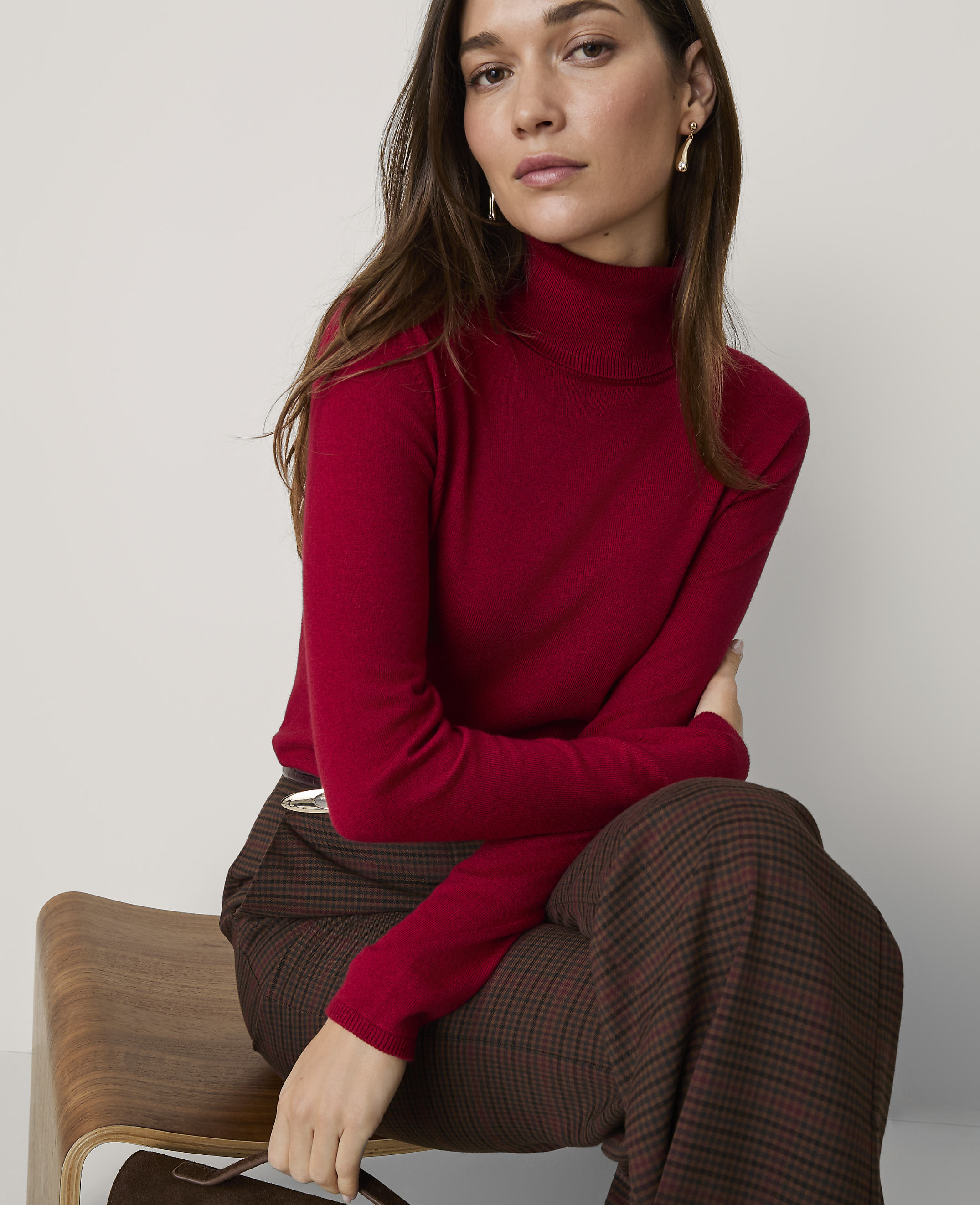A woman wearing a red turtleneck and plaid trousers sits on a wooden chair against a plain white background.