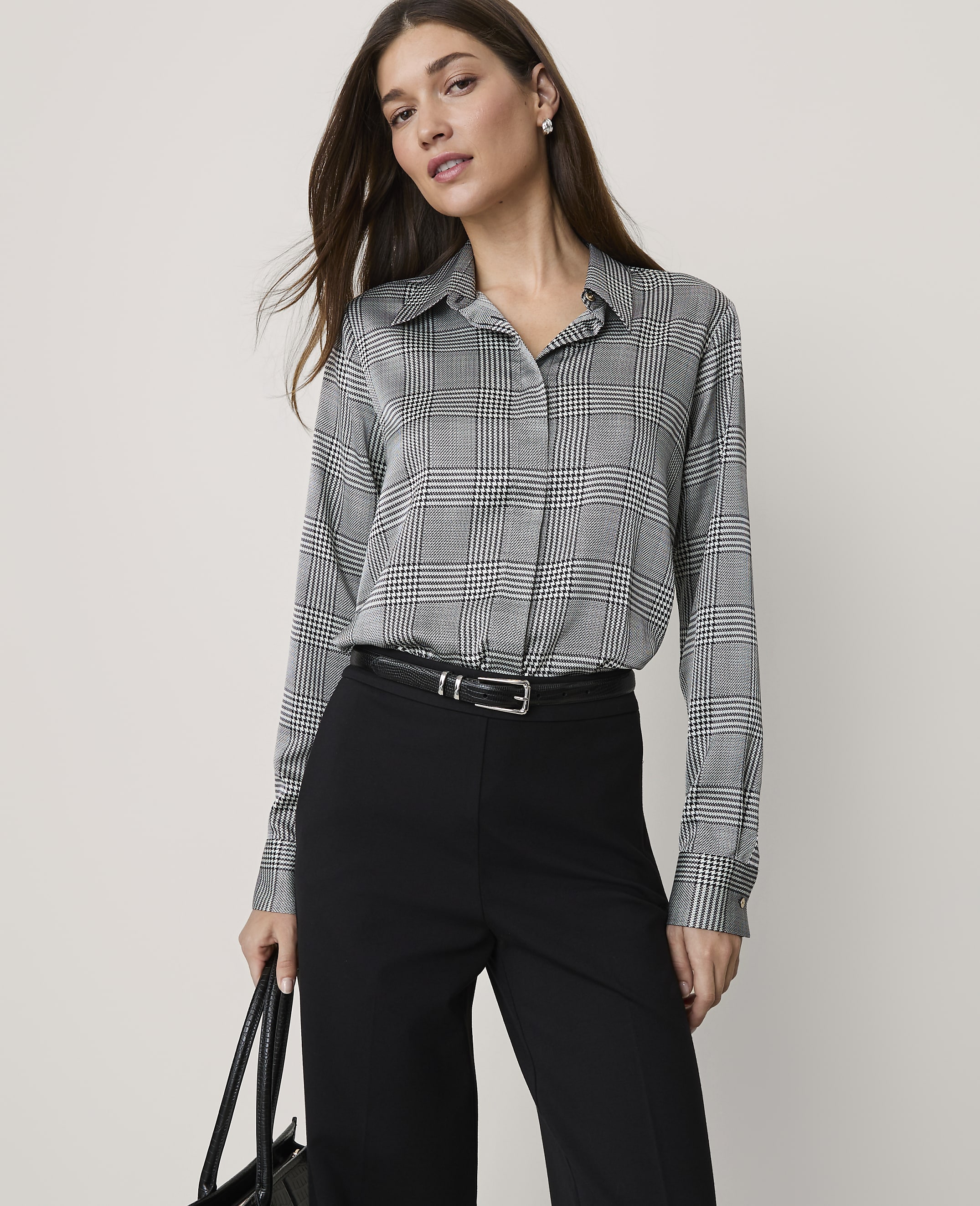 A woman wearing a grey plaid shirt and black trousers stands against a plain, light-colored background.
