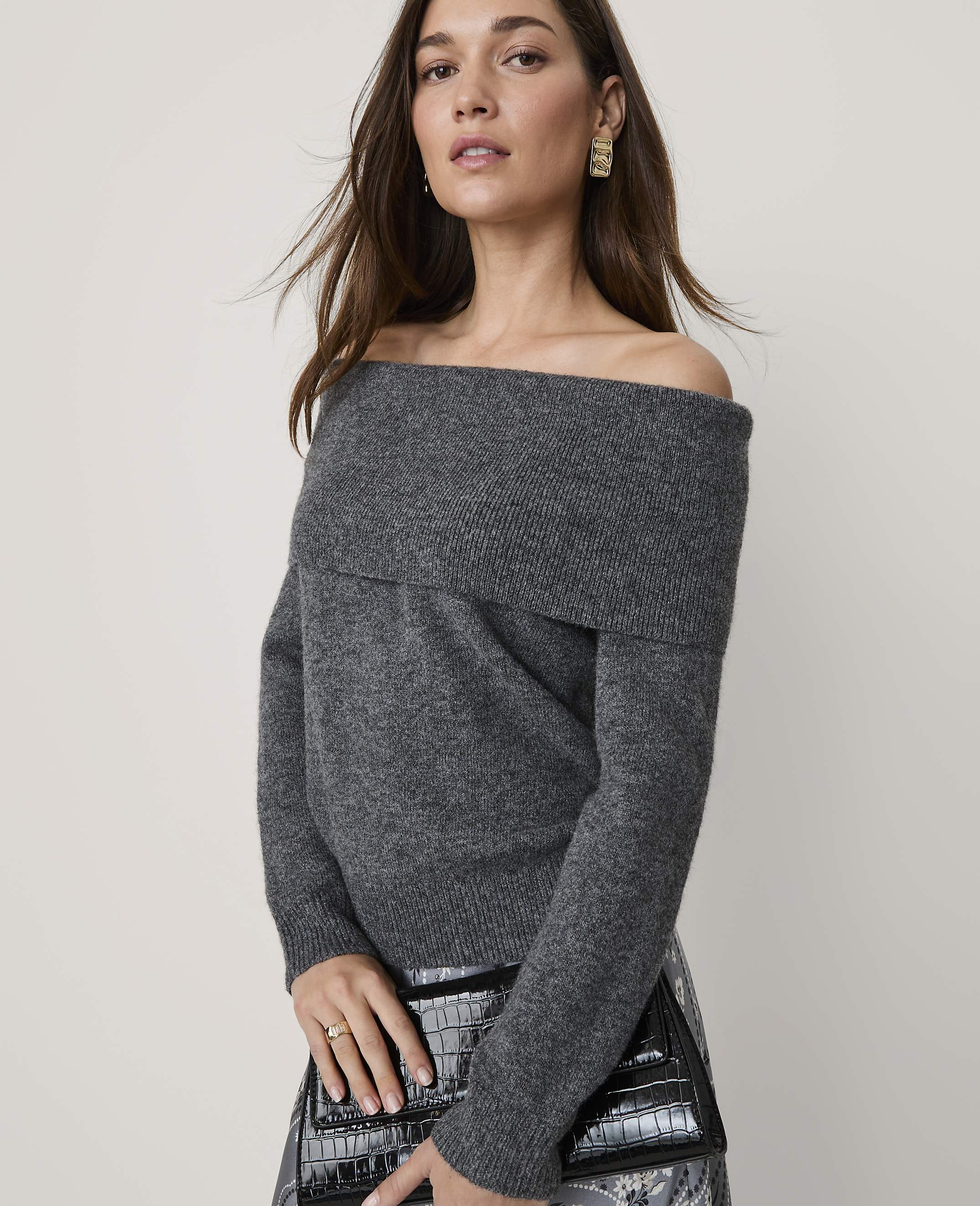 A woman wearing a grey off-the-shoulder sweater and holding a black clutch stands against a plain, light-colored background.
