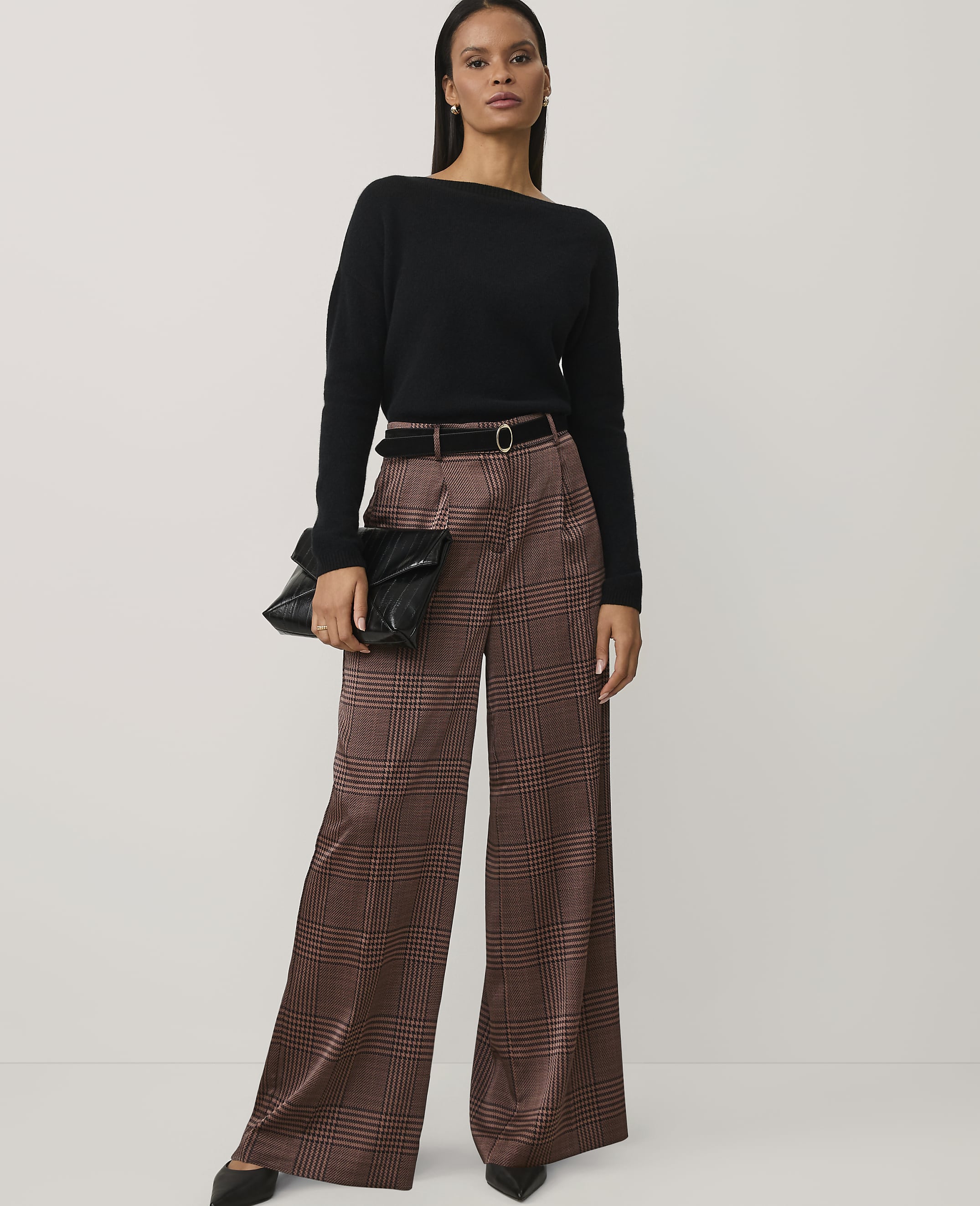 A woman wearing a black sweater and brown plaid wide-leg trousers stands against a plain, light-colored background.