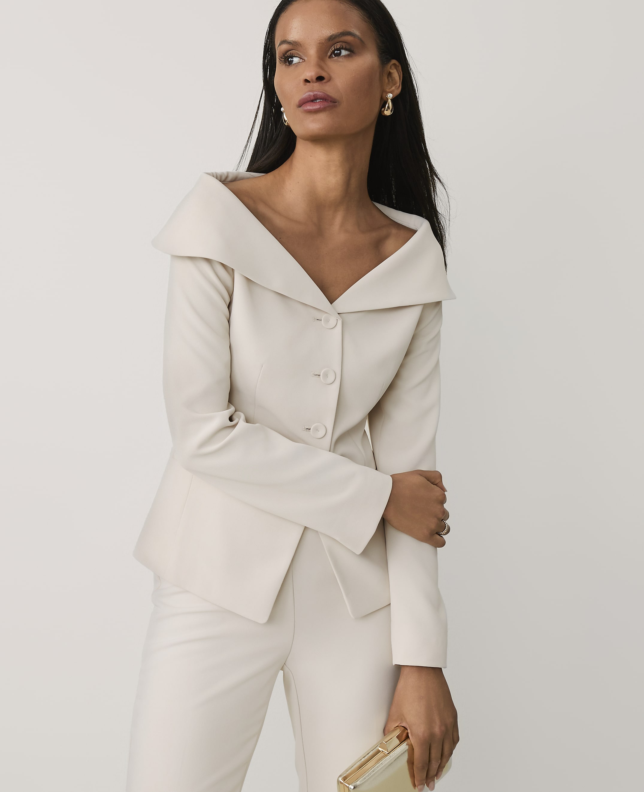 A woman wearing a cream-colored suit and holding a gold clutch stands against a plain white background.