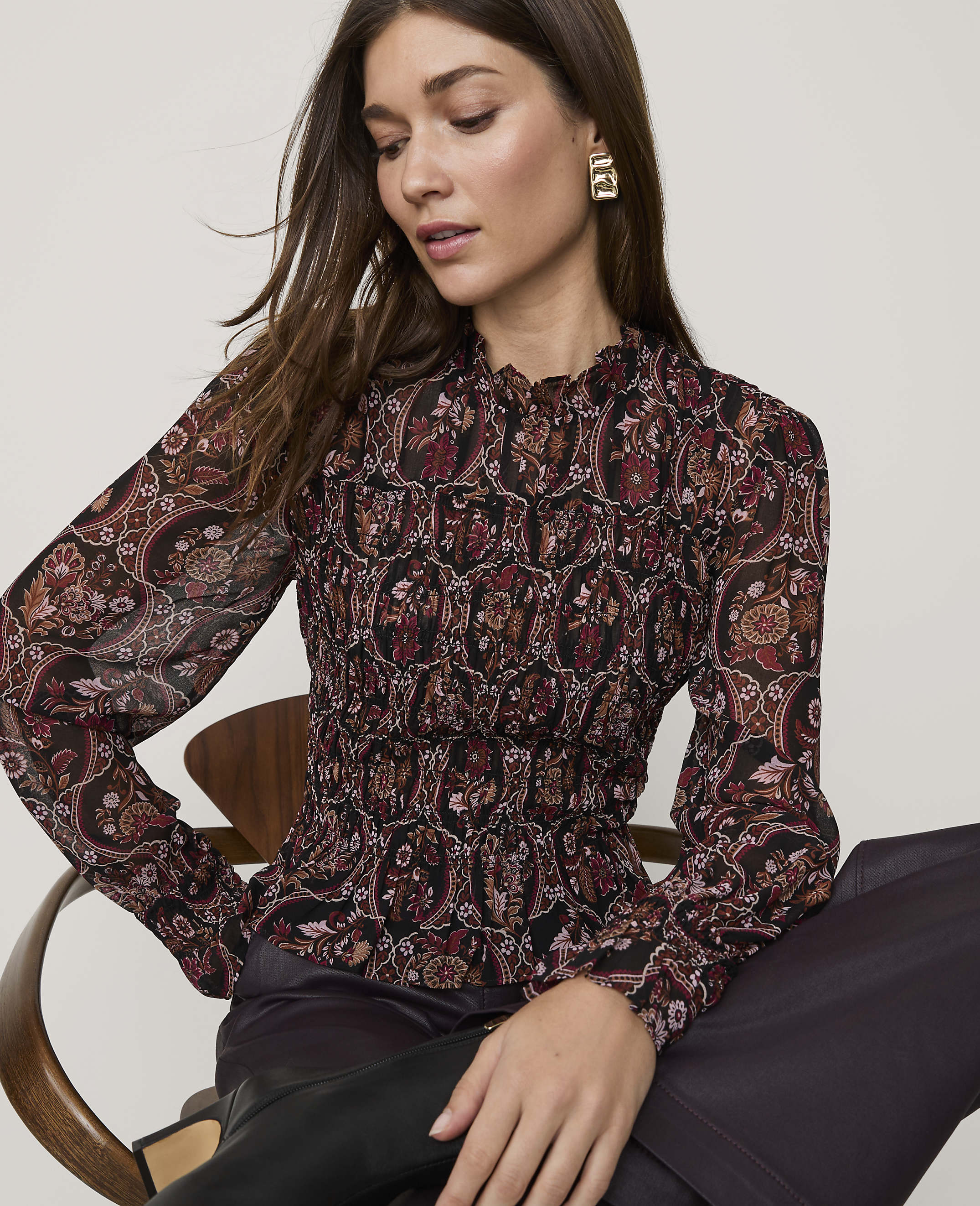 A woman wearing a patterned blouse and dark trousers sits in a wooden chair against a plain white background.